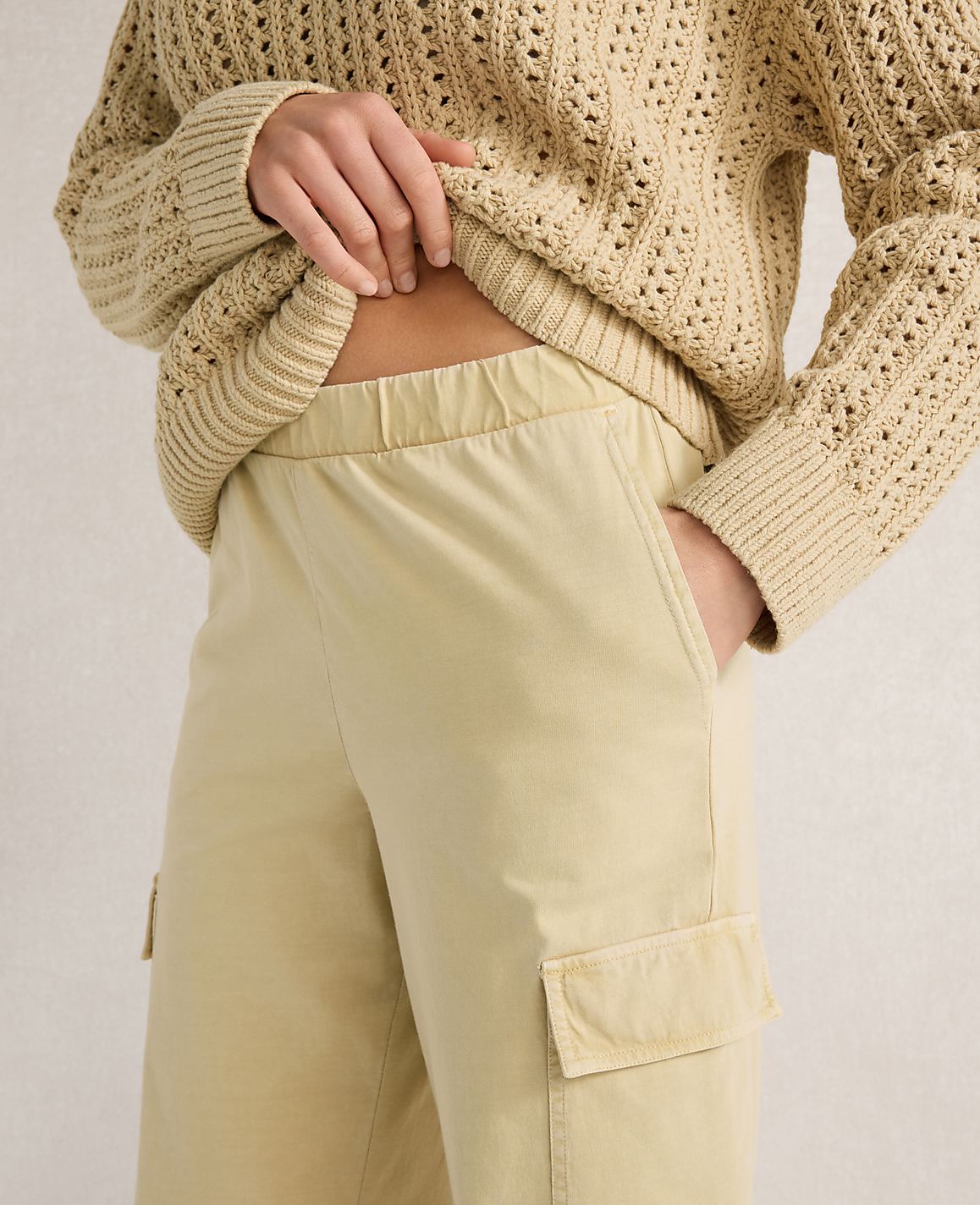 A person in a cream-colored, open-knit sweater and light beige cargo pants stands against a plain, light background.