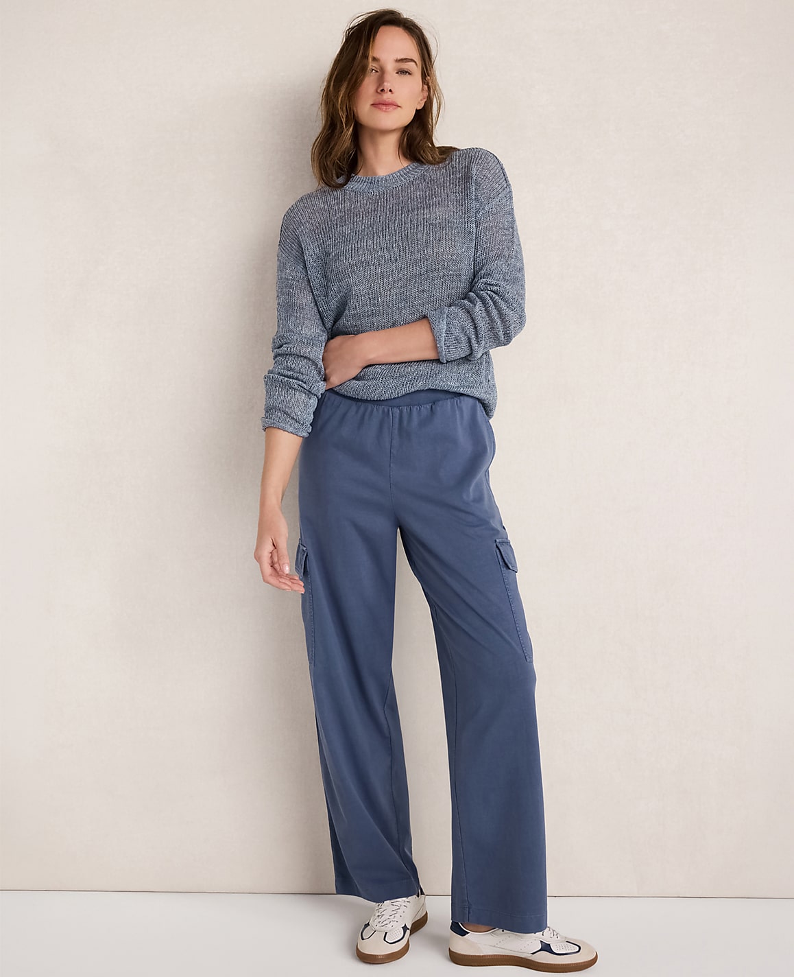 A woman in a blue sweater and blue cargo pants stands against a textured off-white wall.