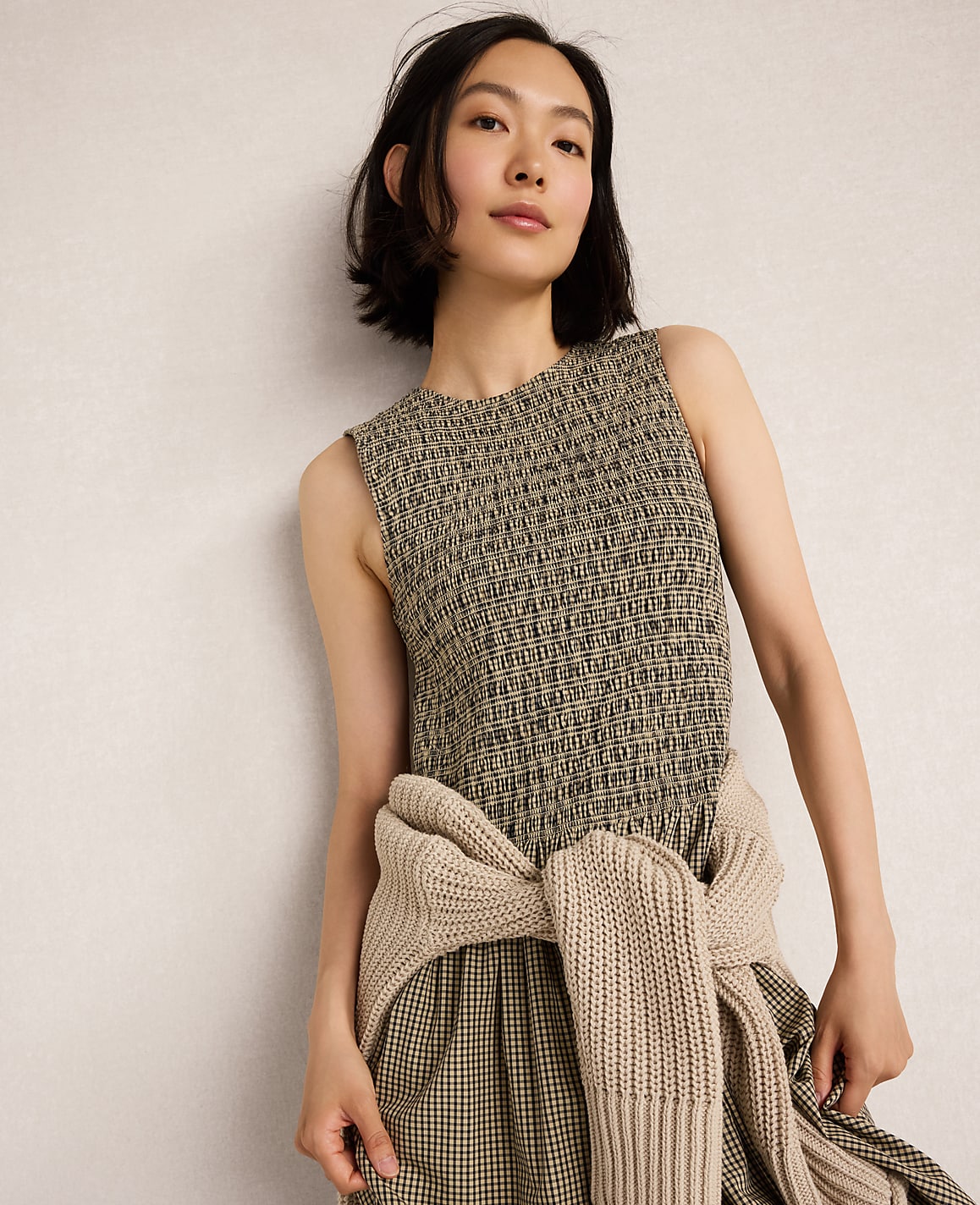 A woman in a textured sleeveless dress and a beige sweater tied around her waist stands against a plain, light-colored wall.