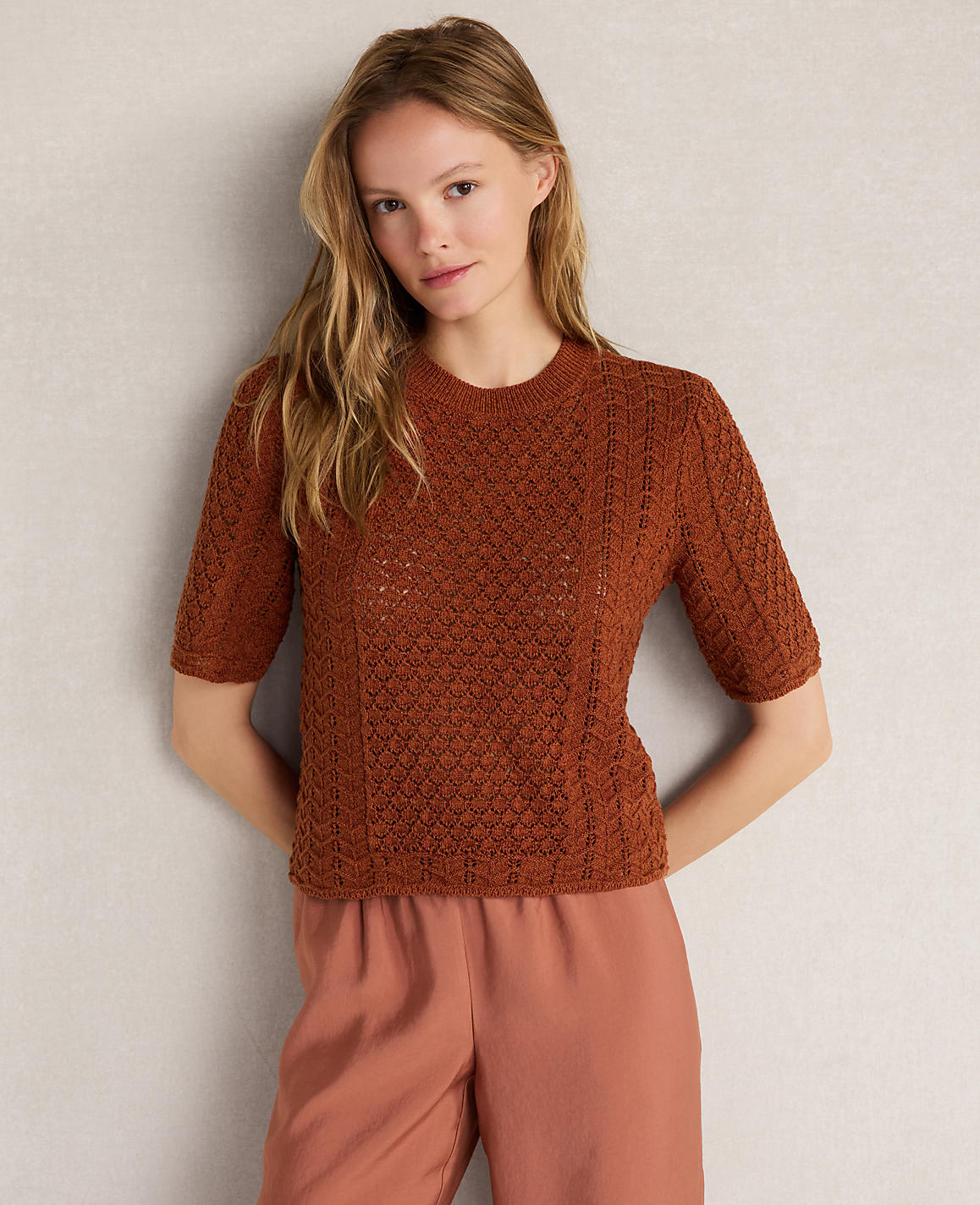 A young woman in a brown knit top and pink pants stands against a plain gray background.