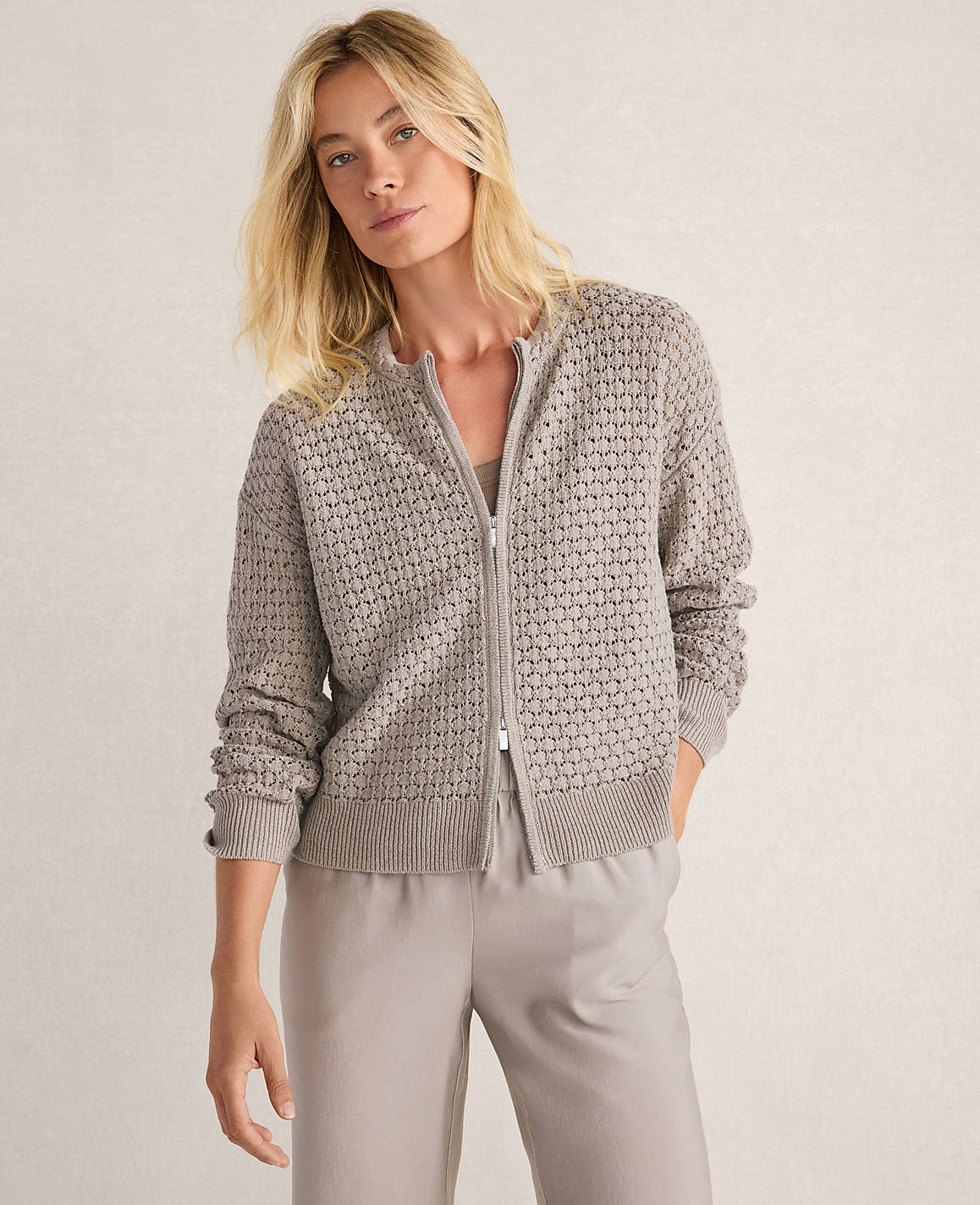 A blonde woman in a textured gray zip-up cardigan and light gray pants stands against a neutral, textured background.