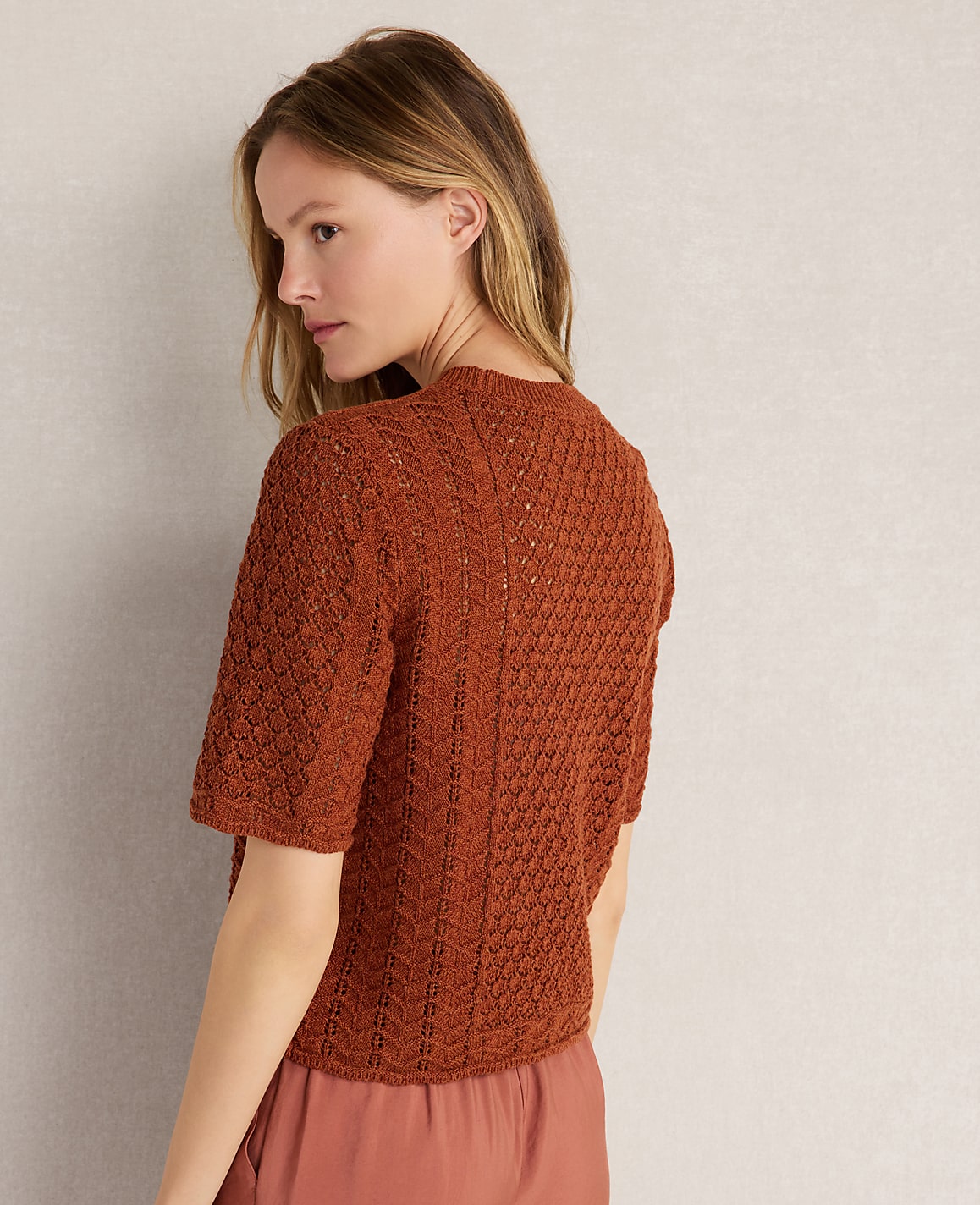 A woman in a rust-colored knit top stands against a plain, light-colored wall.