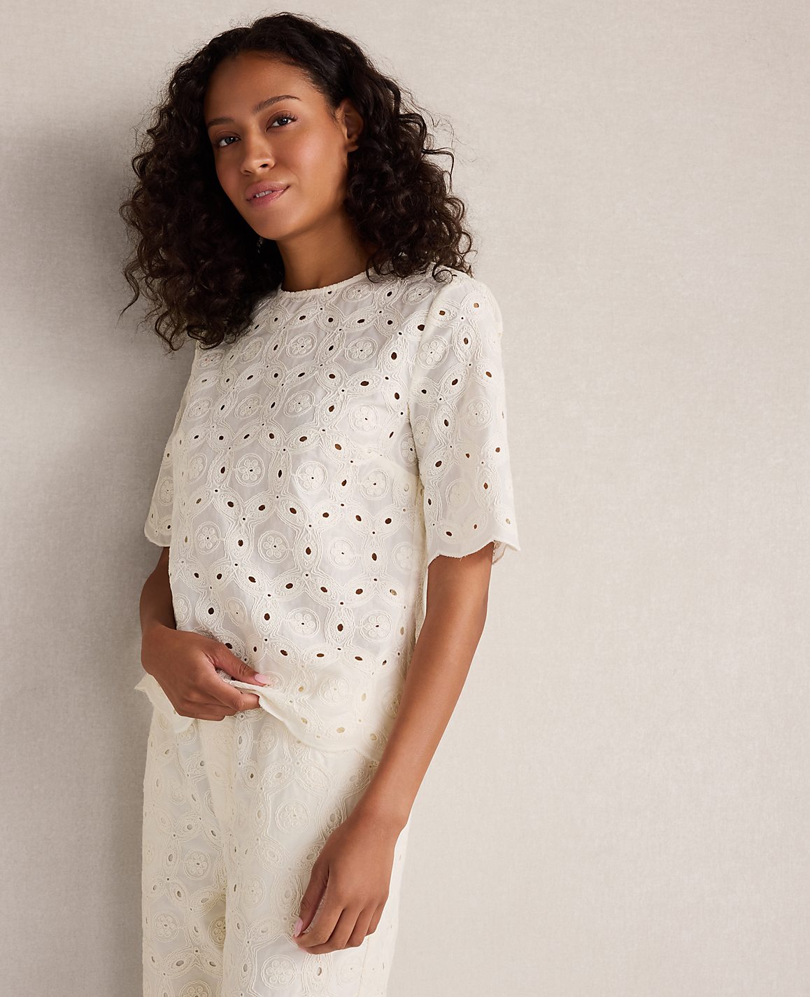 A woman in a white eyelet top and matching pants stands against a plain, textured wall.