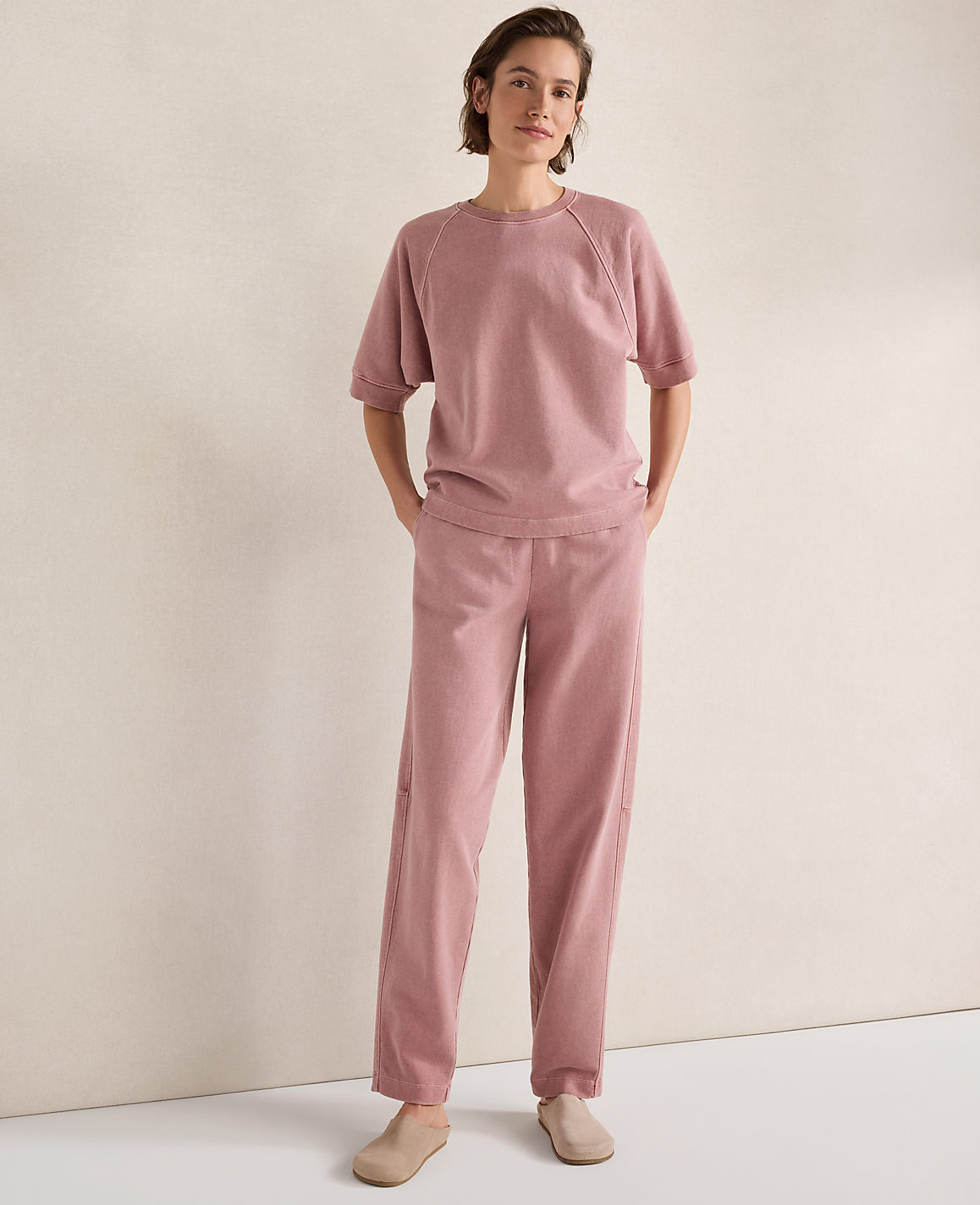 A woman in a pink loungewear set stands with her hands in her pockets against a plain, light-colored wall.