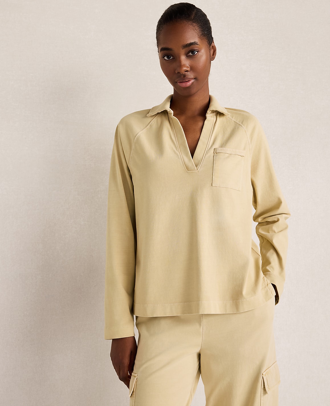 A woman in a beige collared shirt and pants stands against a plain, light-colored background.