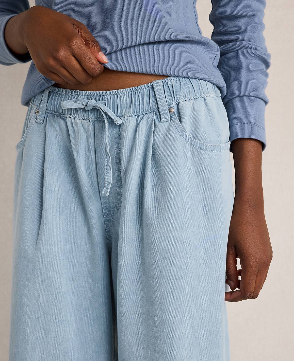 A person's hands adjust the drawstring waist of light blue, wide-leg pants against a plain background.