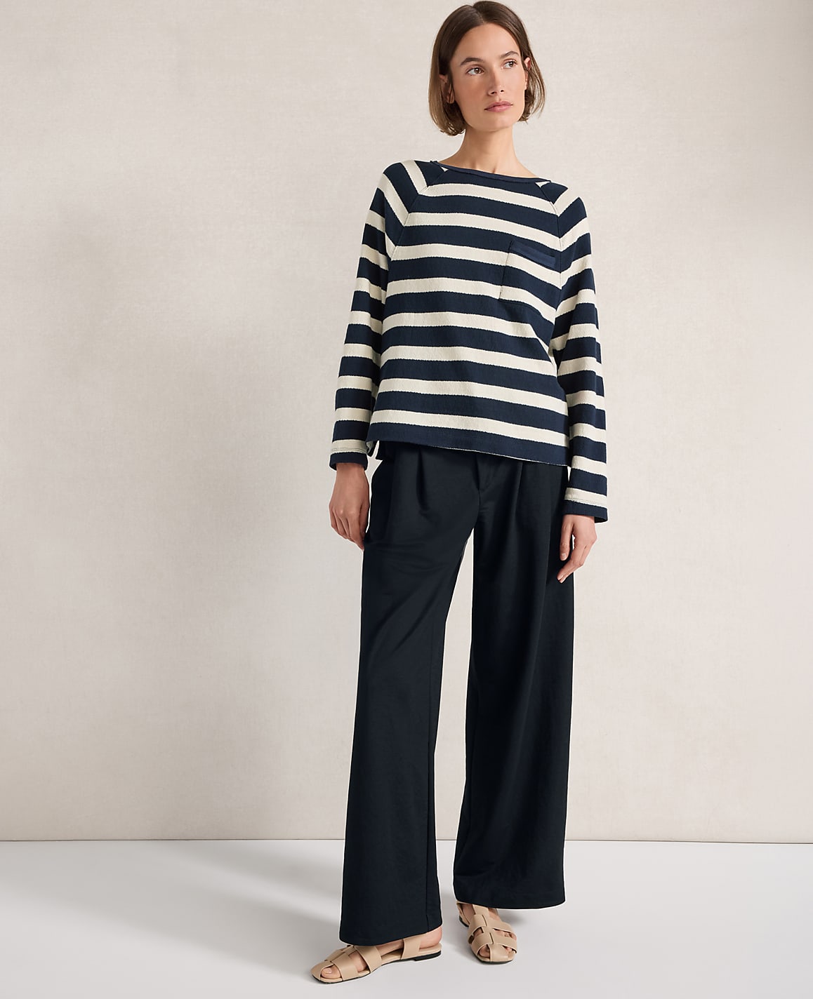 A woman in a striped sweater and wide-leg pants stands against a plain, textured wall.