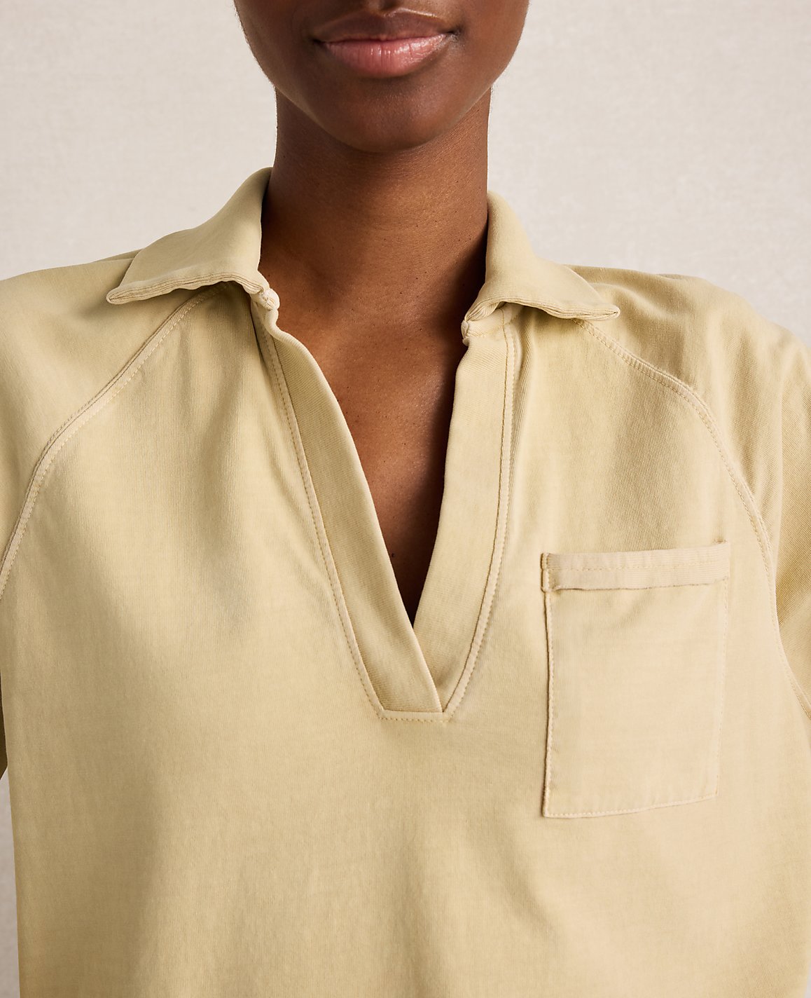 A close-up of a woman wearing a light yellow collared shirt with a pocket, against a plain, light background.