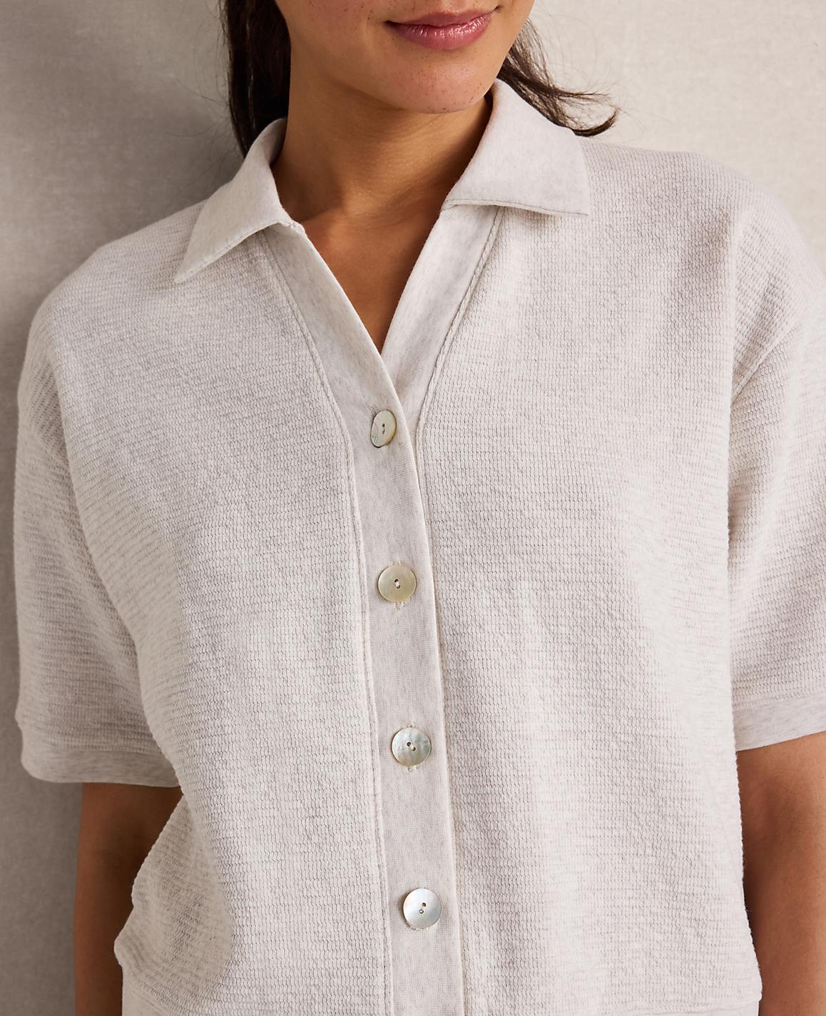 A woman in a textured cream collared shirt with pearl buttons is shown against a neutral, textured background.
