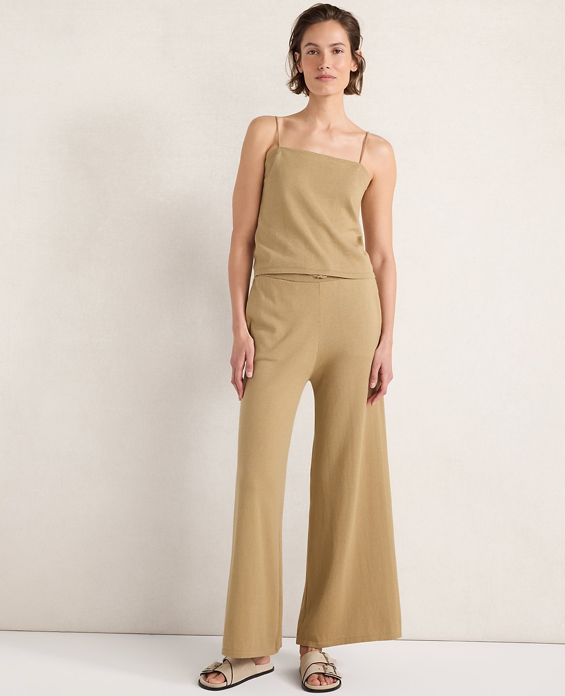 A woman in a tan knit camisole and wide-leg pants stands against a plain, textured cream background.