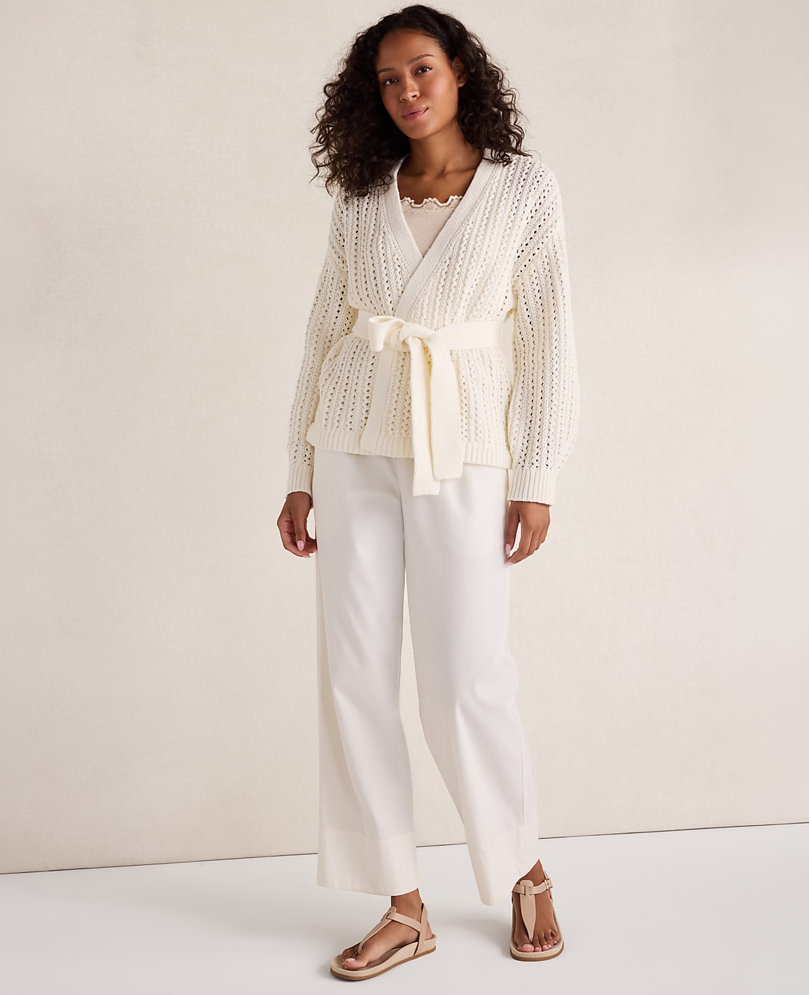 A woman in a cream-colored wrap cardigan and wide-leg pants stands against a plain, light-colored background.