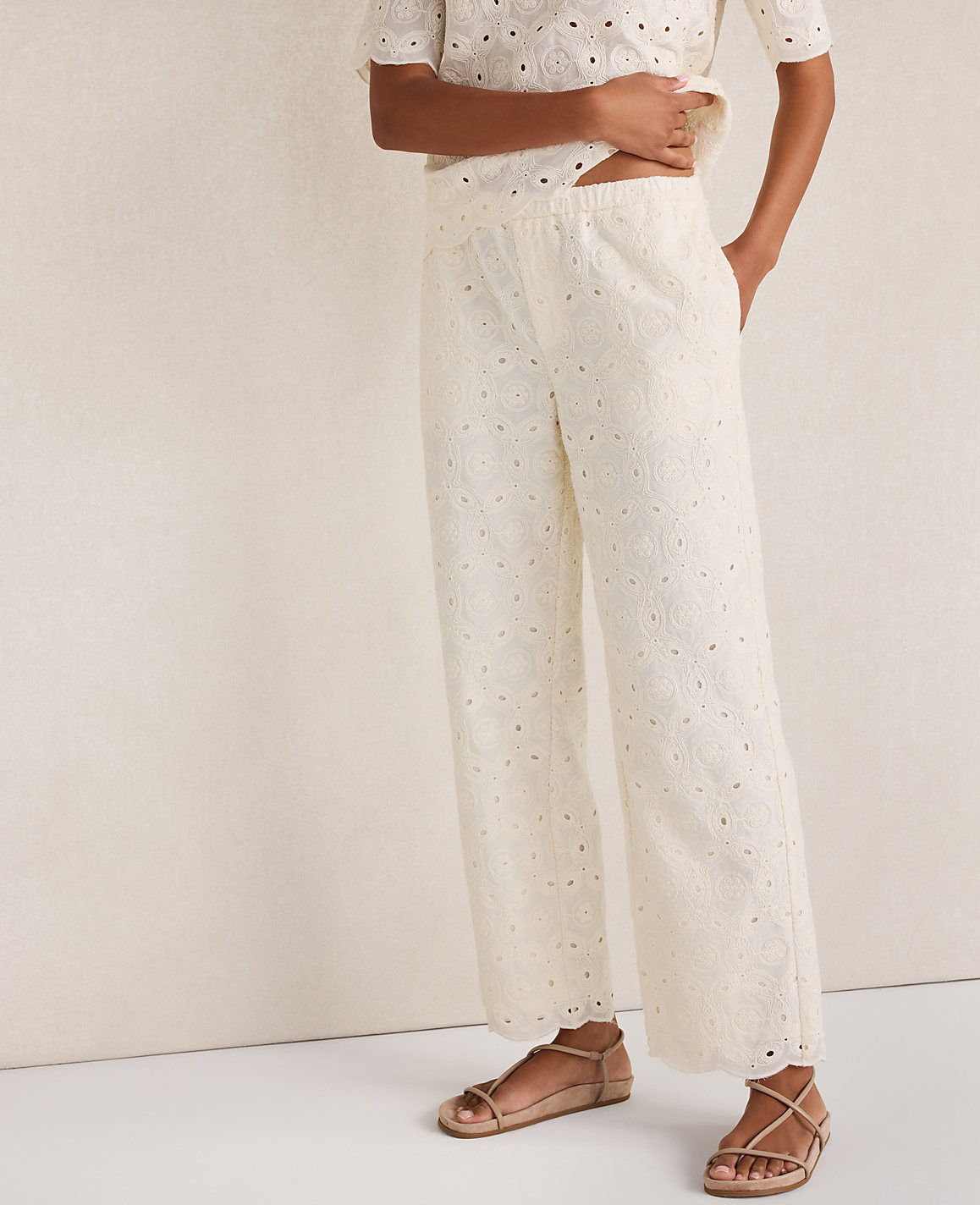 A person stands in cream eyelet pants and sandals against a plain, light-colored background.