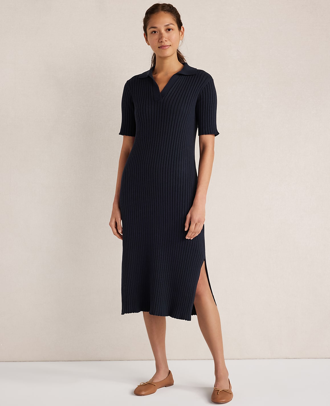 A woman in a navy ribbed polo dress and brown flats stands against a plain, light-colored background.