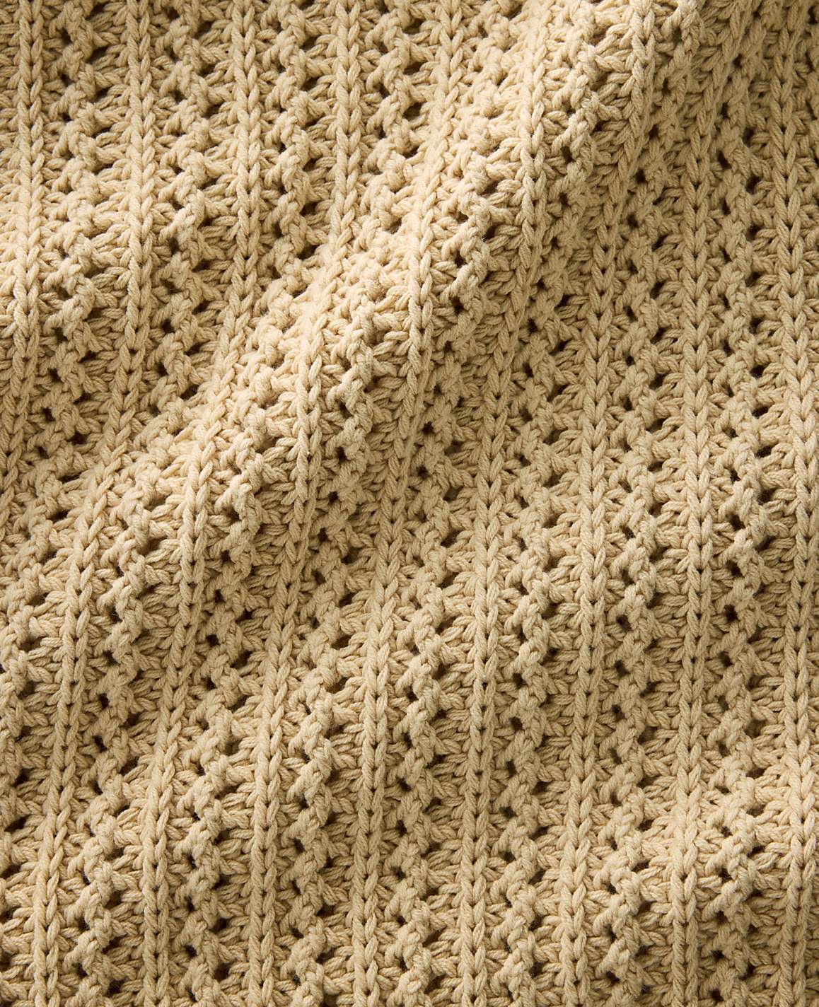 A textured, beige crocheted fabric with a repeating openwork pattern is draped and folded.