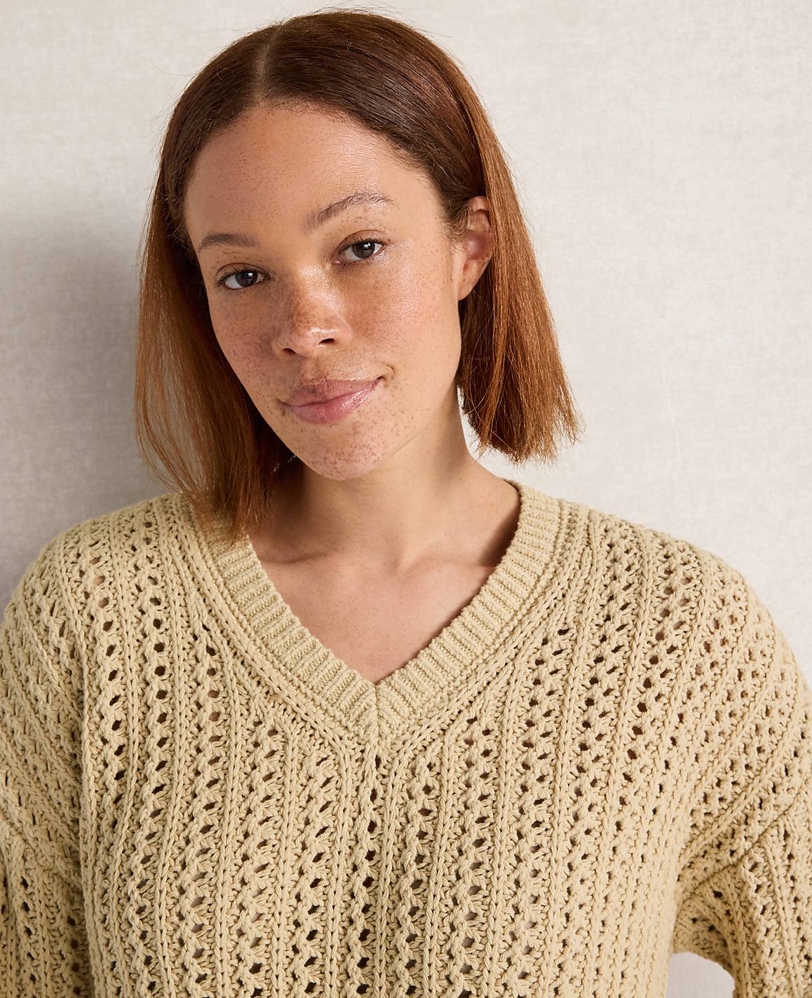 A woman with short brown hair and freckles smiles gently in a light yellow, open-knit sweater against a textured, light-colored background.