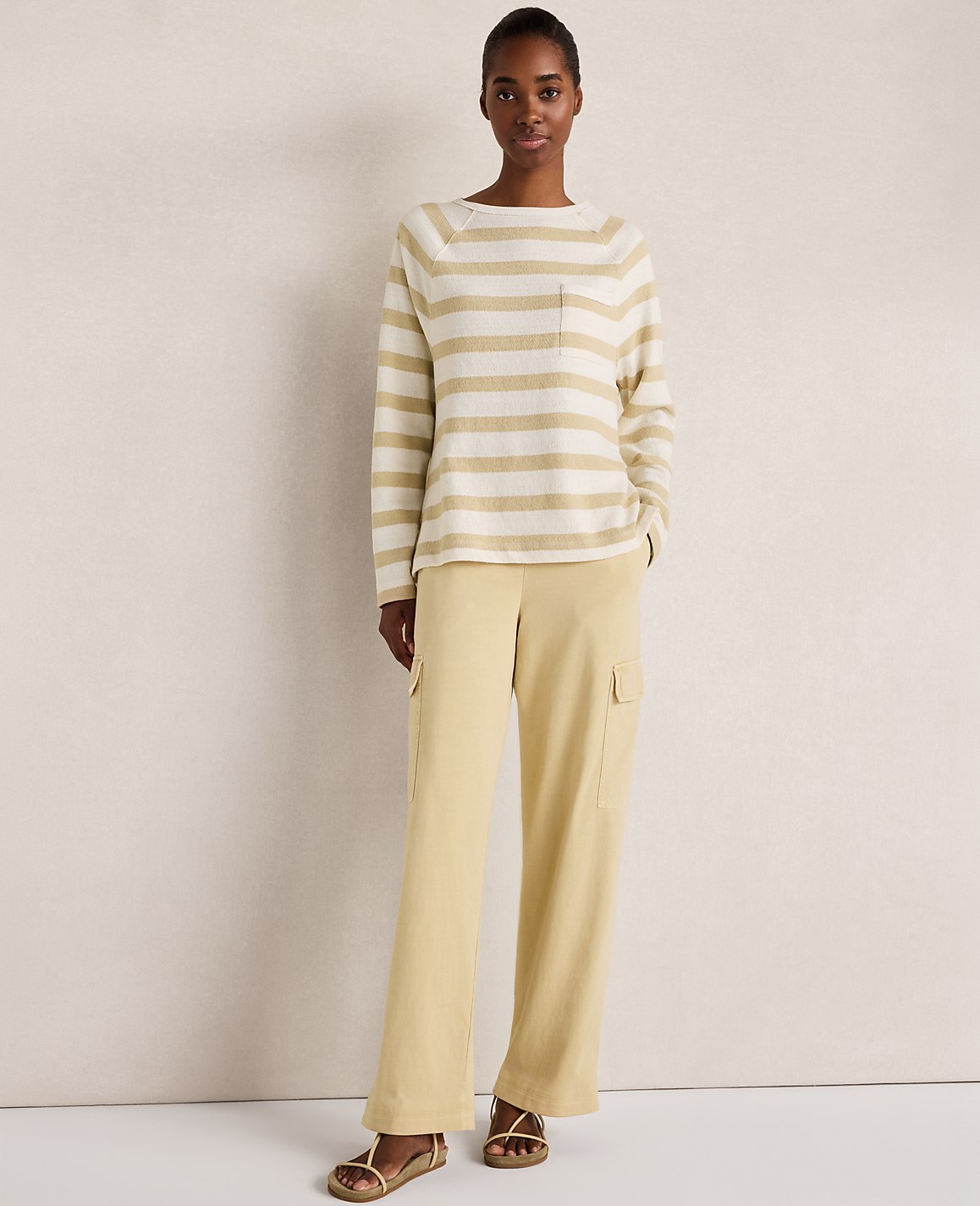 A woman in a striped sweater and yellow pants stands against a textured, light-colored wall.