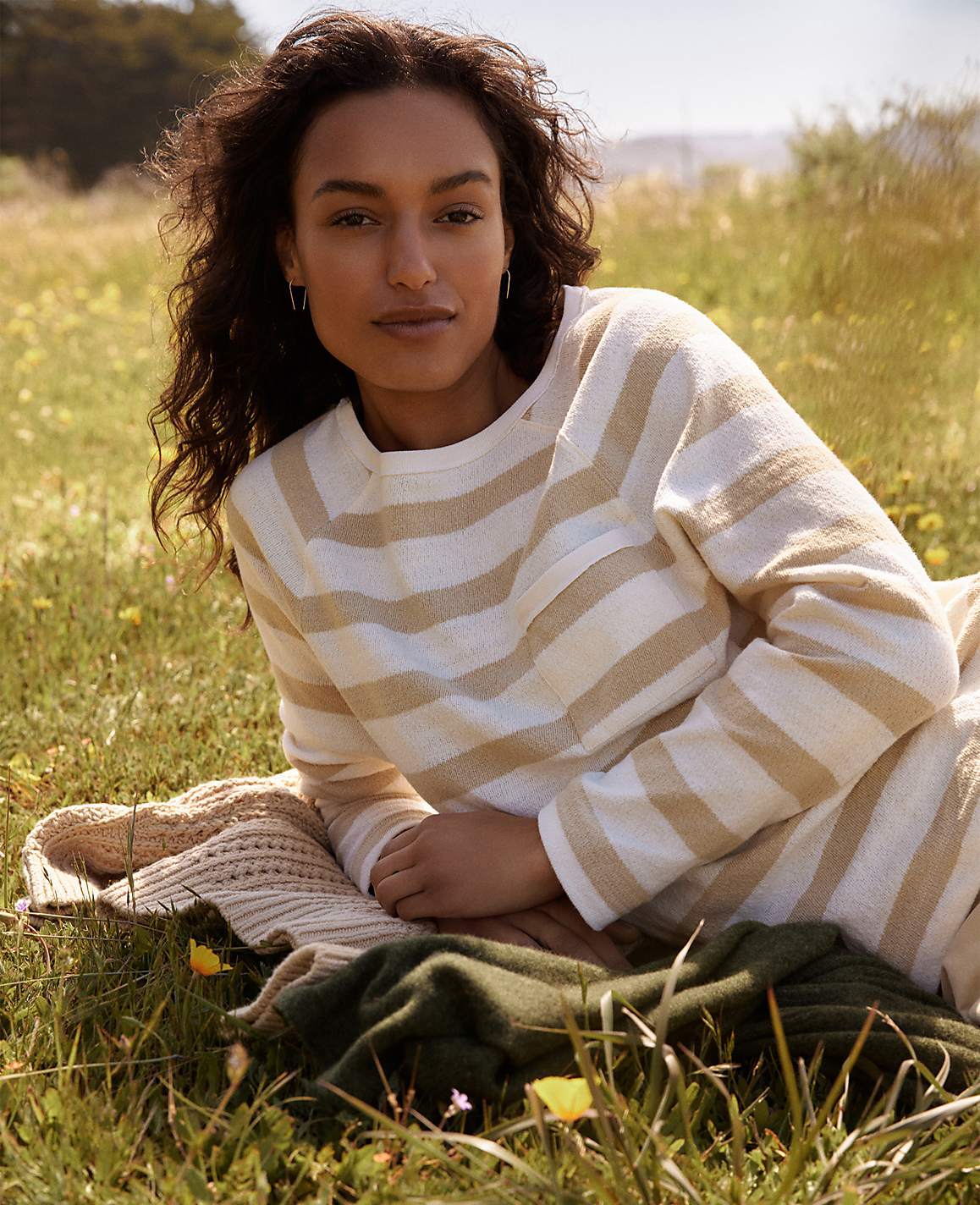 A woman with curly dark hair lies on a blanket in a grassy field with yellow wildflowers, while a blurred landscape stretches out behind her.