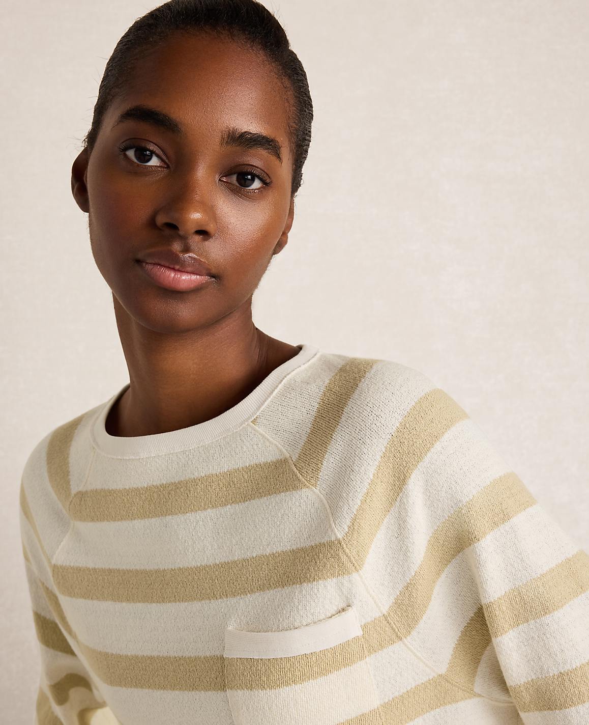 A woman with dark skin and short hair wears a cream and tan striped sweater with a pocket in the foreground, against a softly lit, neutral background.