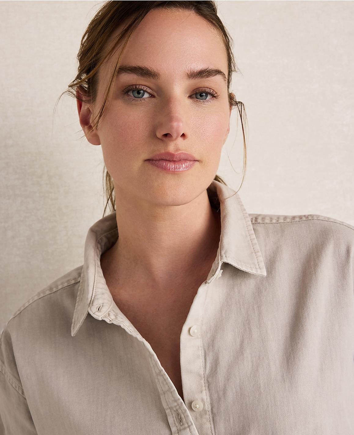 A woman with light skin and blue eyes, wearing a light-colored button-down shirt, is captured in a close-up shot against a textured, neutral background.