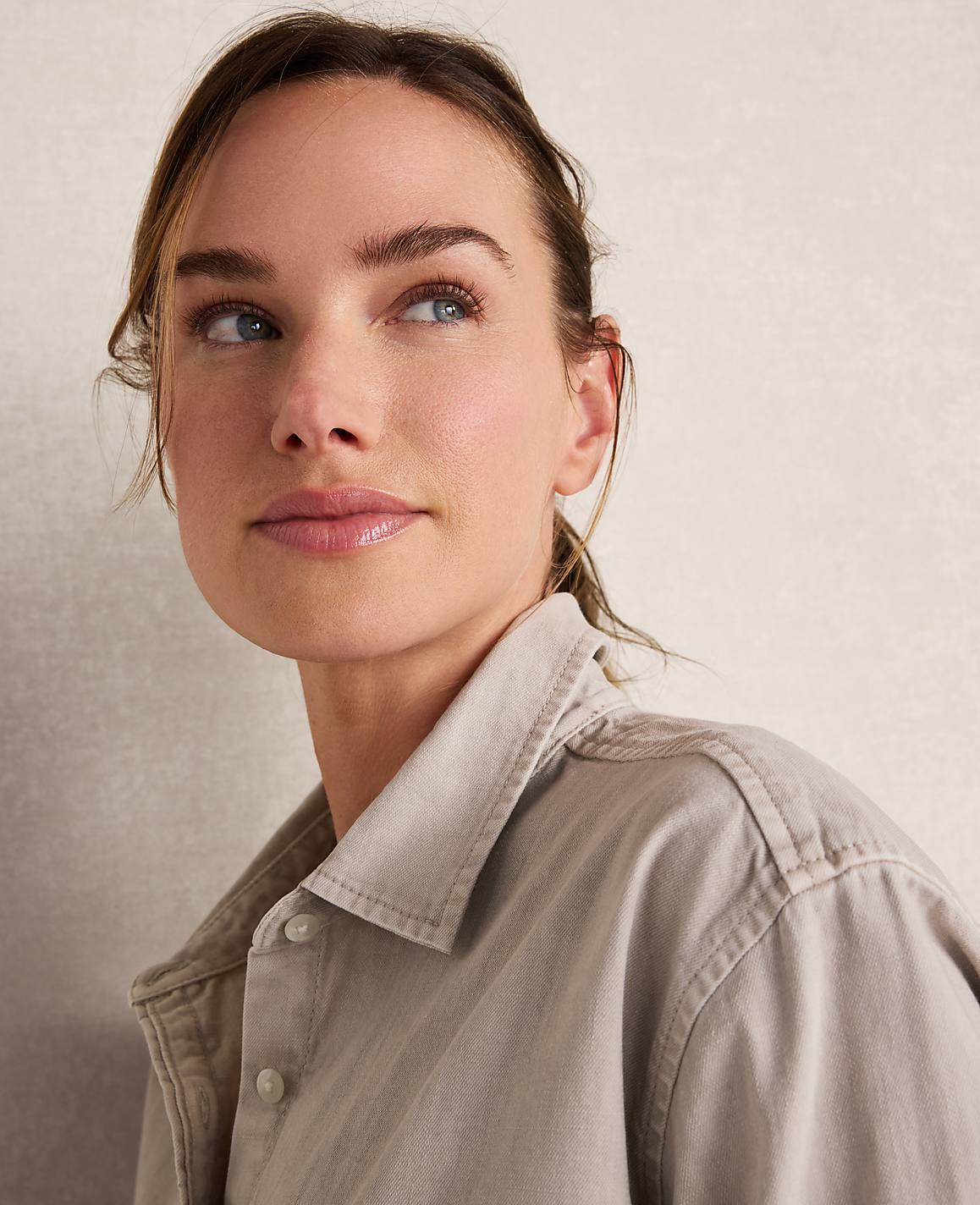 A woman with light skin and brown hair pulled back looks up and to the left, wearing a light brown collared shirt against a plain, textured background.