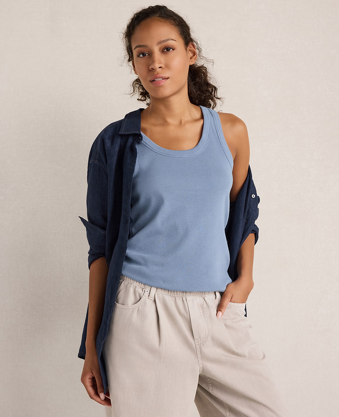 A woman in a blue tank top and tan pants stands against a plain, light-colored background.