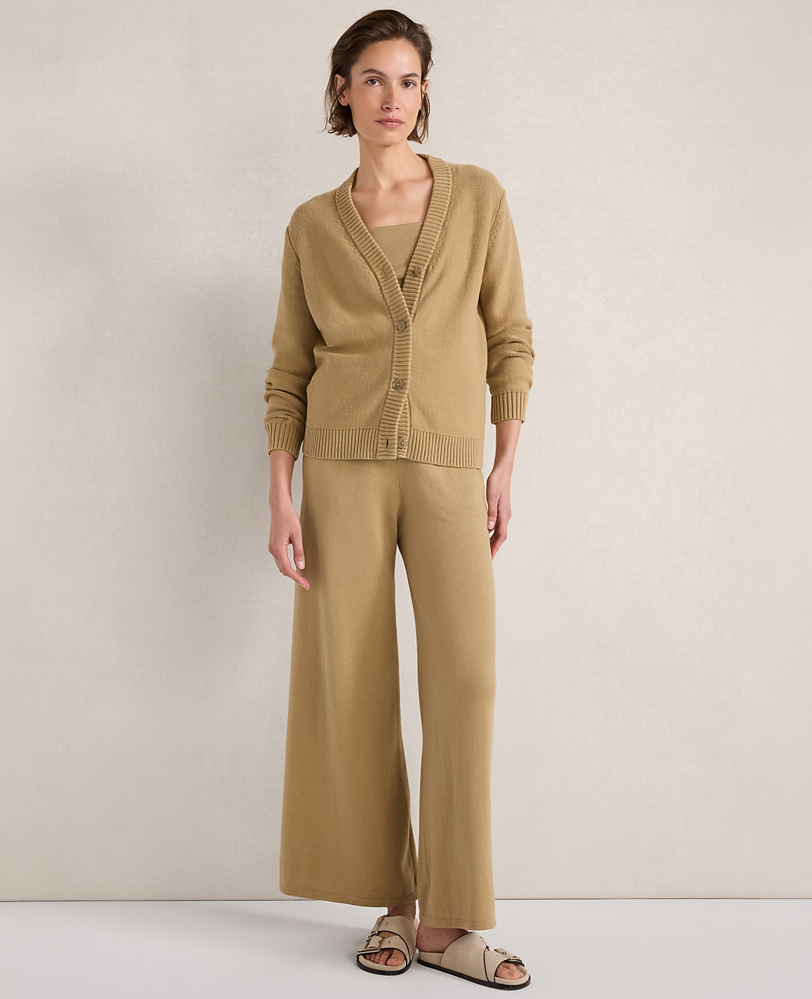 A woman in a beige cardigan, tank top, and wide-leg pants stands against a plain, textured wall.