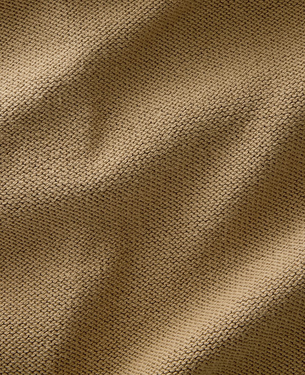 A close-up view of a textured, tan knitted fabric fills the frame, with soft shadows and highlights creating depth across its surface.