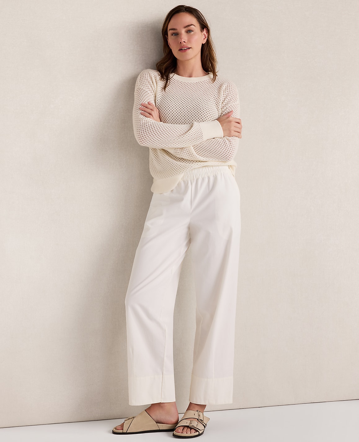 A woman in a cream sweater and white pants stands with her arms crossed against a textured cream wall.