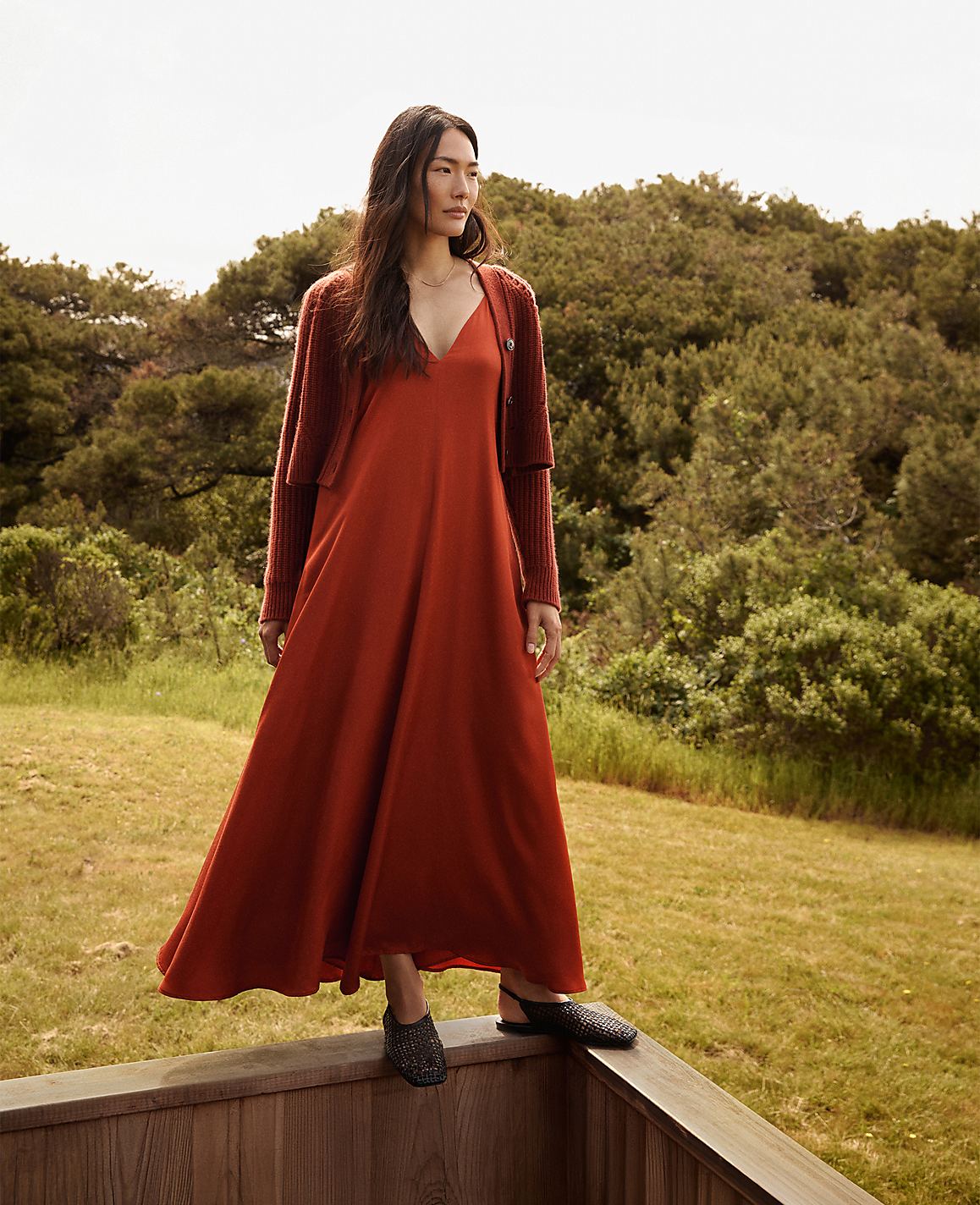 A woman in a rust-colored dress and cardigan stands on a wooden railing with a lush green forest in the background.