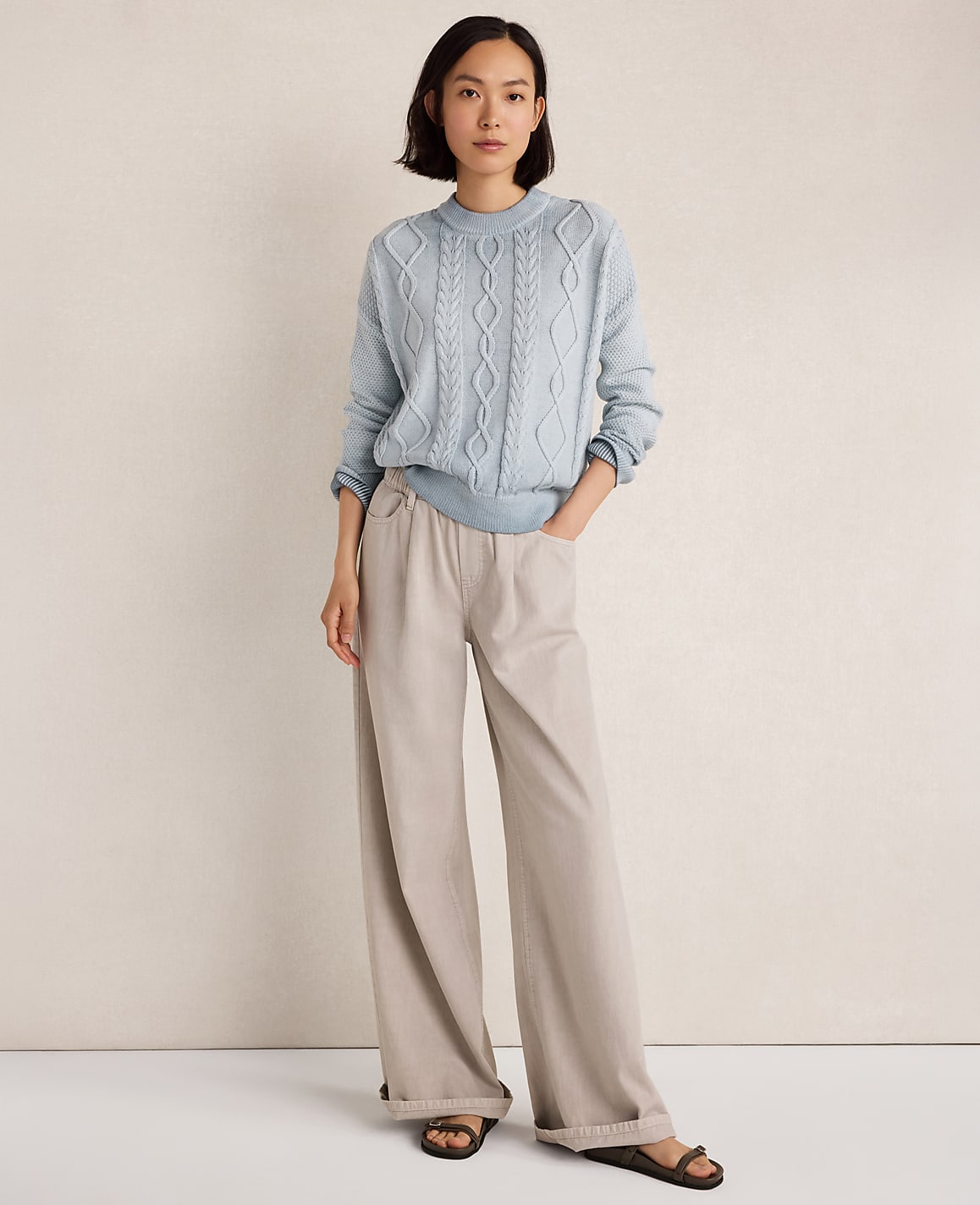 A woman in a light blue cable-knit sweater and beige wide-leg pants stands against a textured off-white wall.