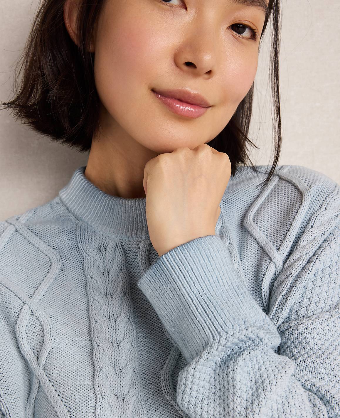A young woman with short dark hair rests her chin on her hand, wearing a light blue cable-knit sweater against a soft, textured background.