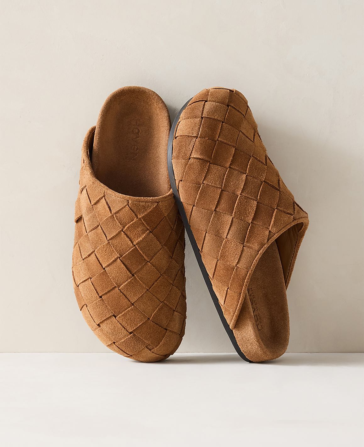 A pair of tan woven suede clogs rests on a light-colored surface against a plain, light background.