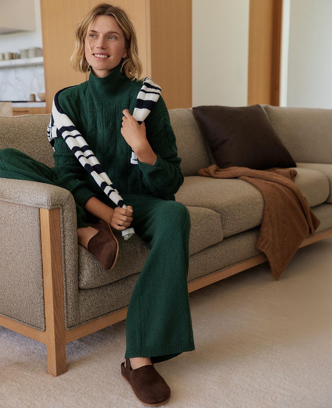 A woman in a green sweater and pants sits on a couch with a striped scarf draped over her shoulder, while a blurred background shows a modern living room.