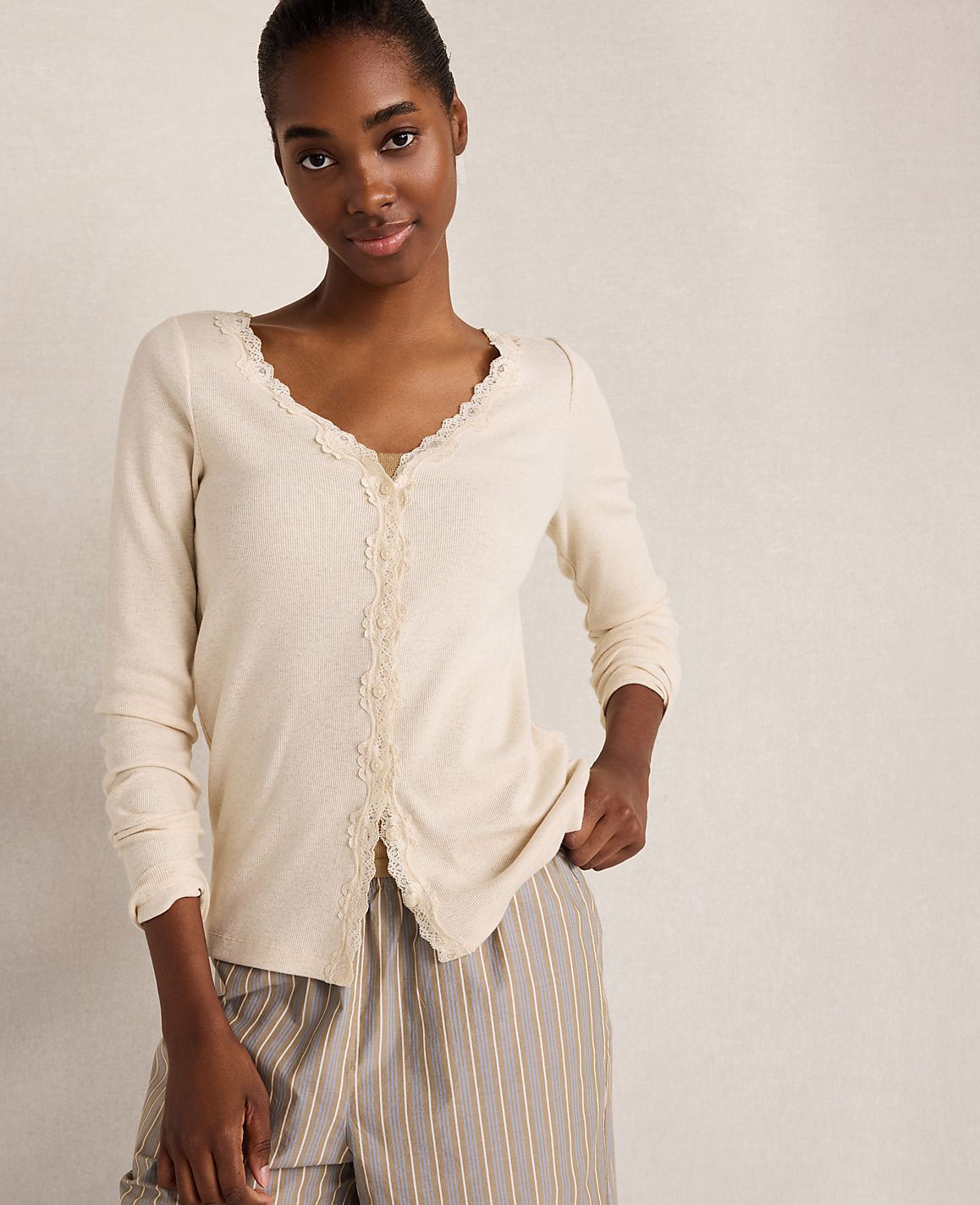 A woman in a cream-colored lace-trimmed top and striped pants stands against a plain, light-colored background.