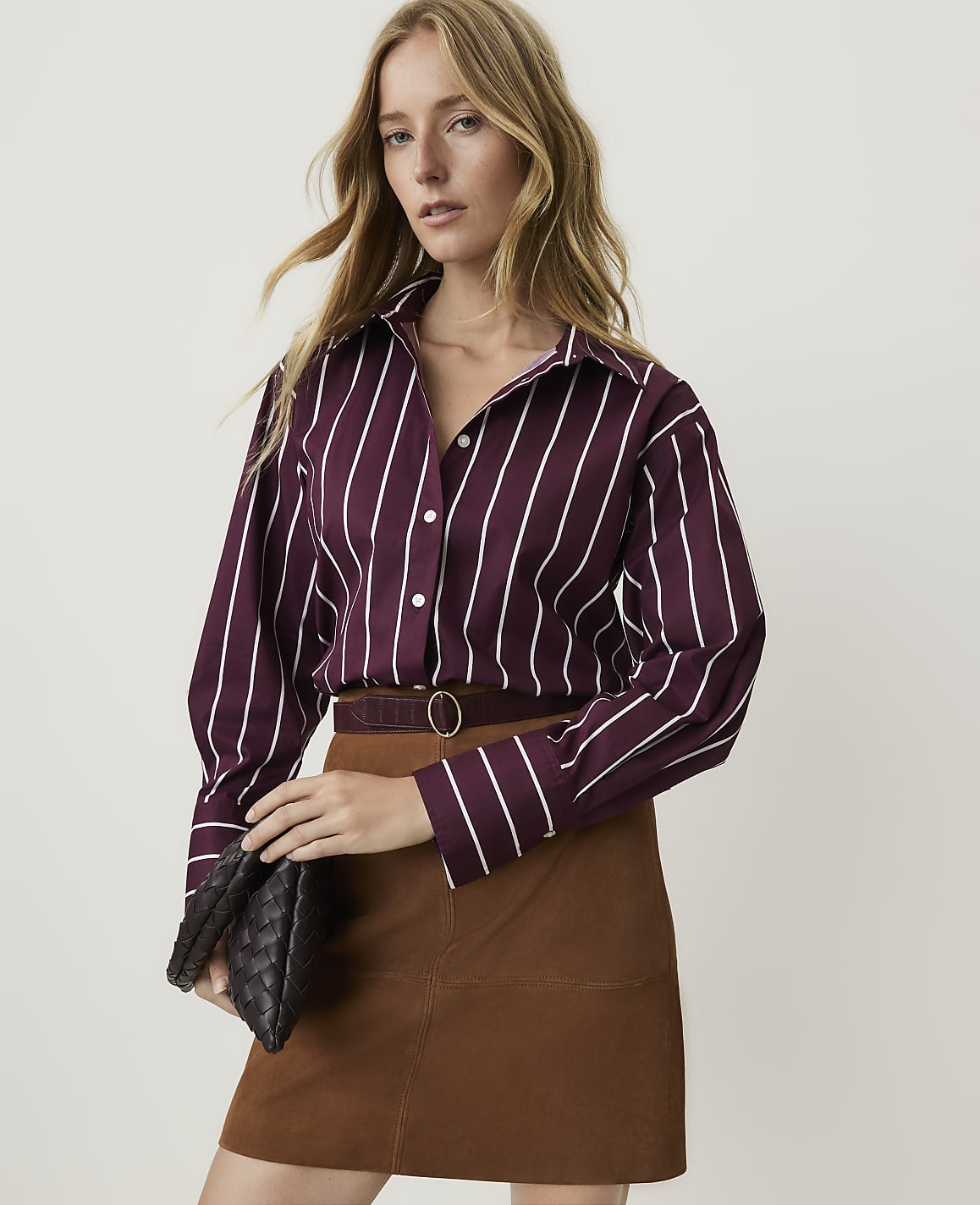 A woman in a striped shirt and brown skirt holds a clutch in front of a plain background.
