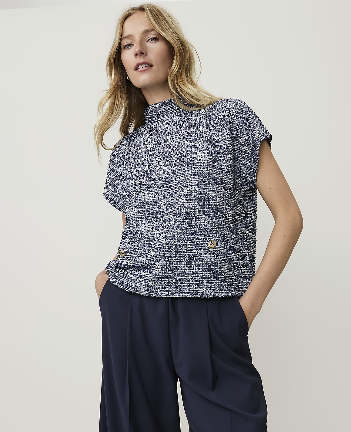 A woman in a textured blue top and dark pants stands against a plain, light gray background.