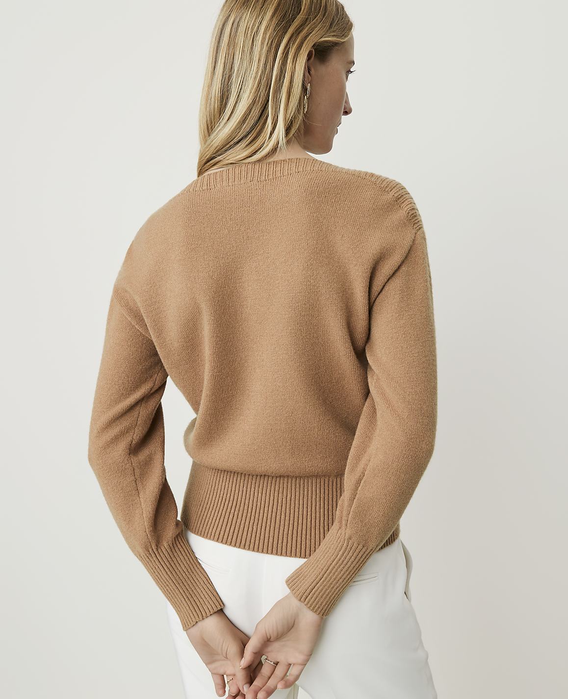 A woman in a tan sweater and white pants stands with her back to the camera against a plain white background.
