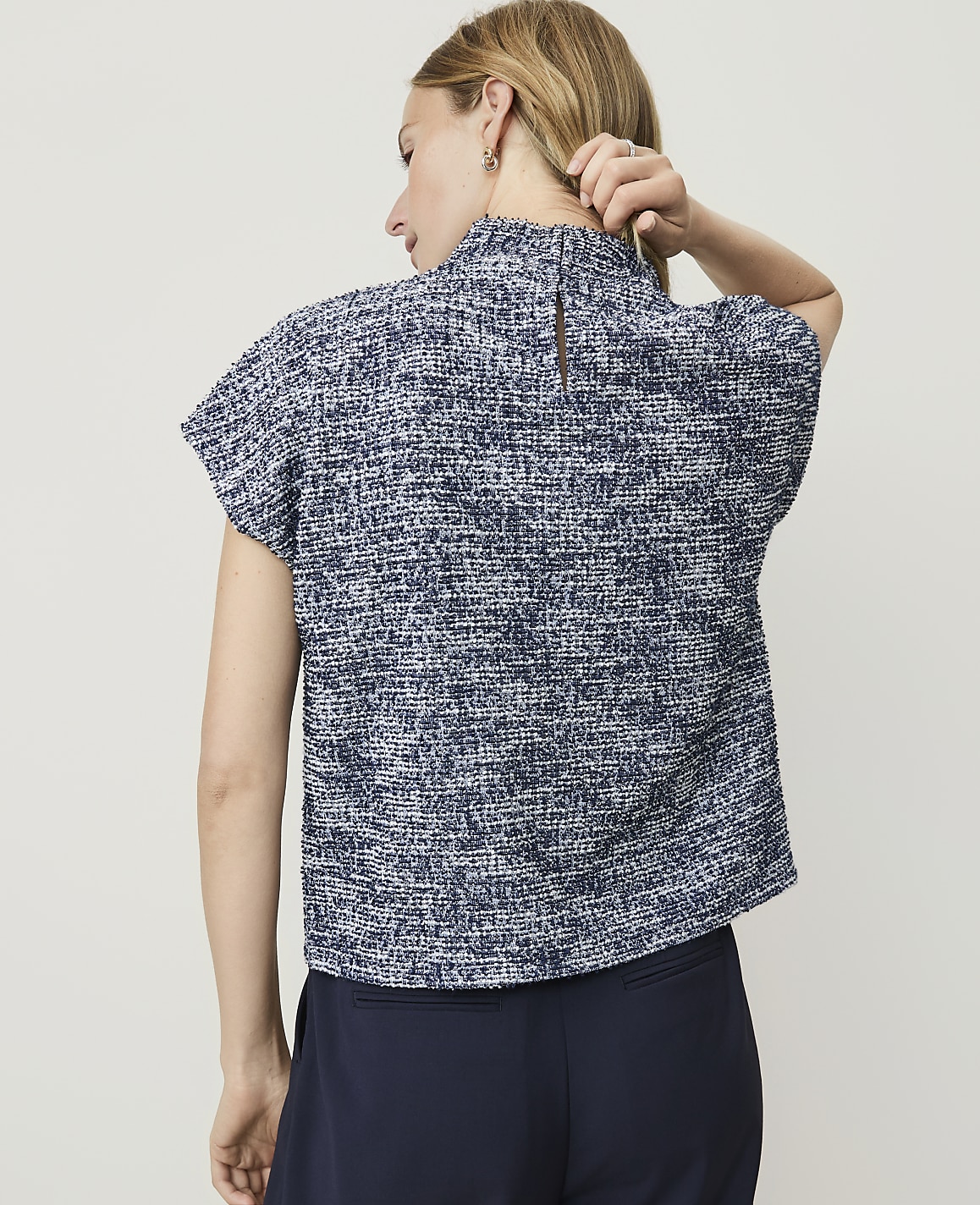 A woman in a textured blue top and dark pants stands against a plain, light background.
