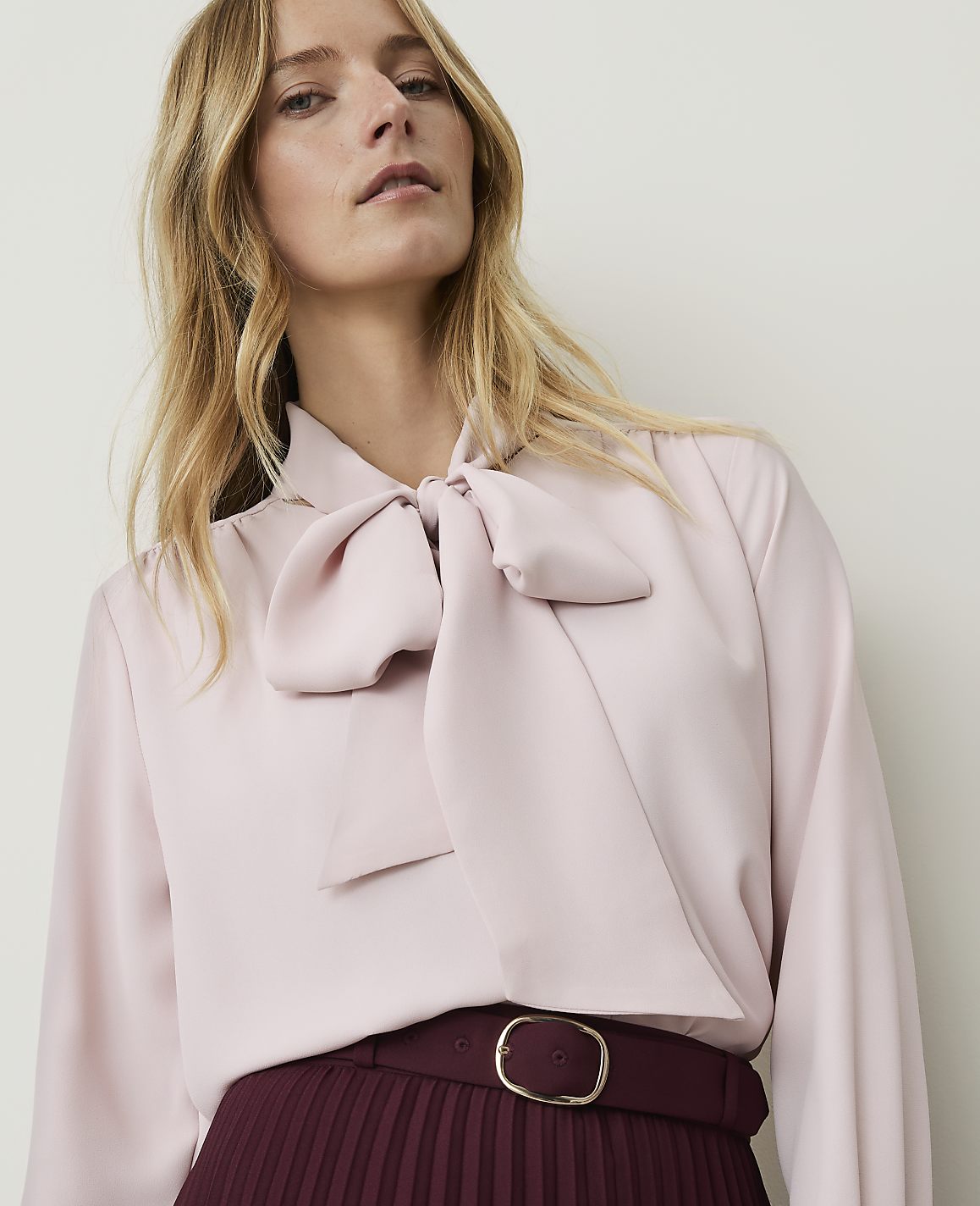 A woman with blonde hair wears a pale pink blouse with a large bow at the neck and a burgundy pleated skirt with a belt, against a plain white background.
