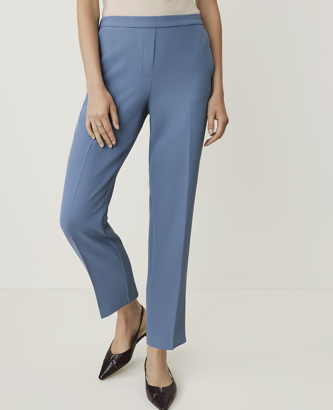 A woman in blue trousers and dark pointed shoes stands against a plain white background.