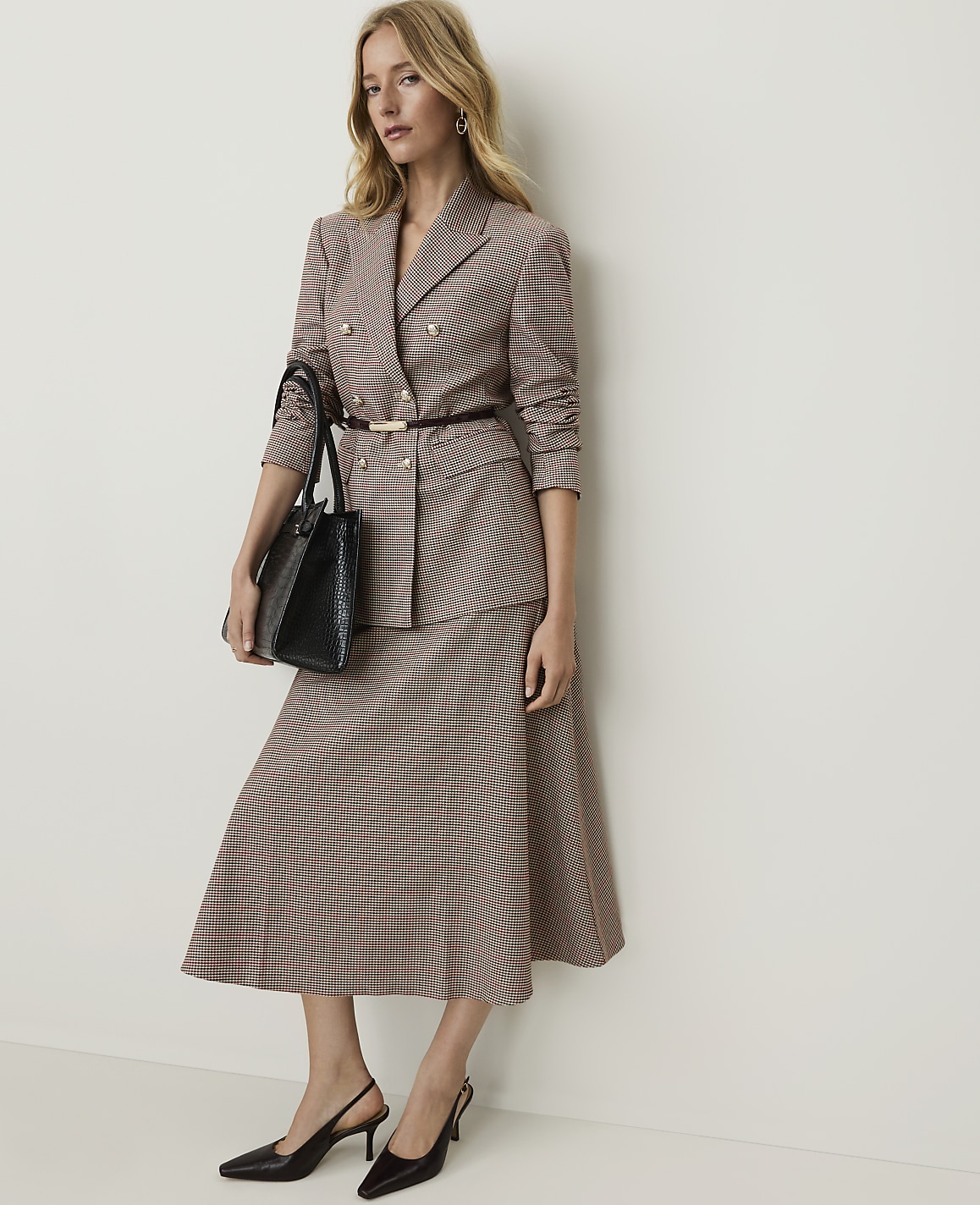 A woman in a patterned suit and heels stands holding a black handbag against a plain, light-colored wall.