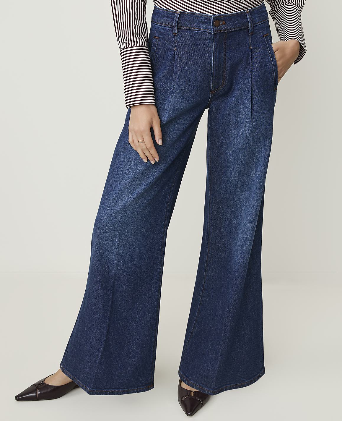 A person in dark wash, wide-leg denim jeans and pointed-toe shoes stands against a plain white background.