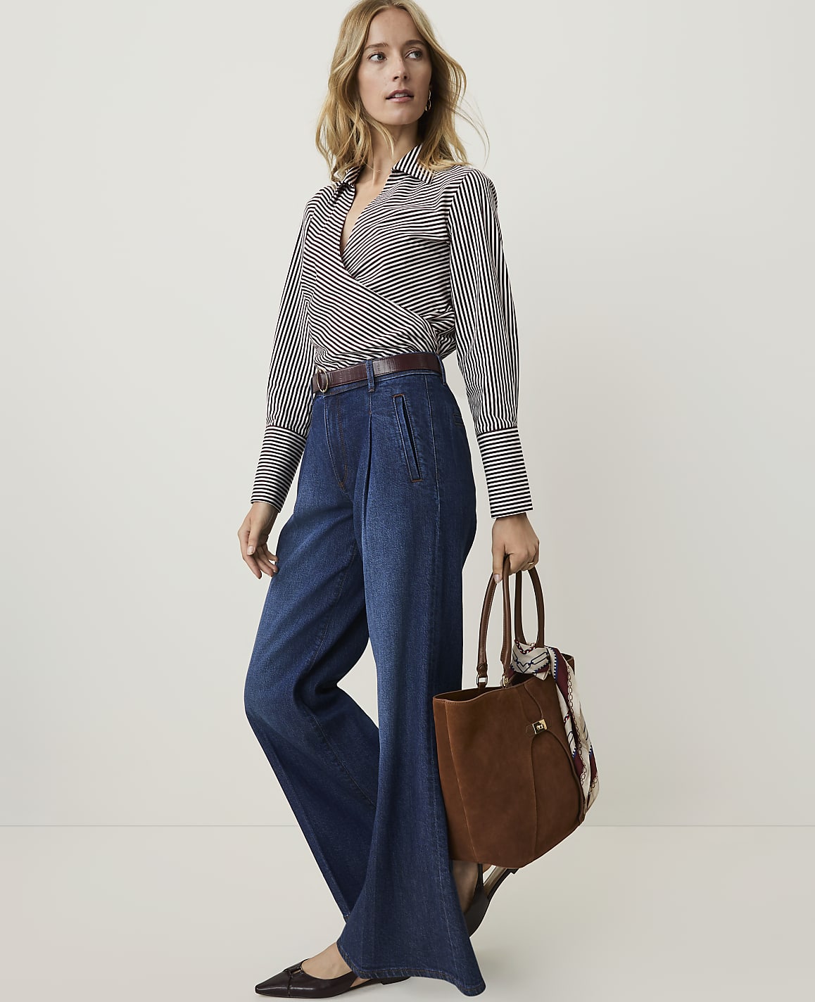 A woman in a striped blouse and wide-leg jeans stands against a plain white background, holding a brown leather tote bag.