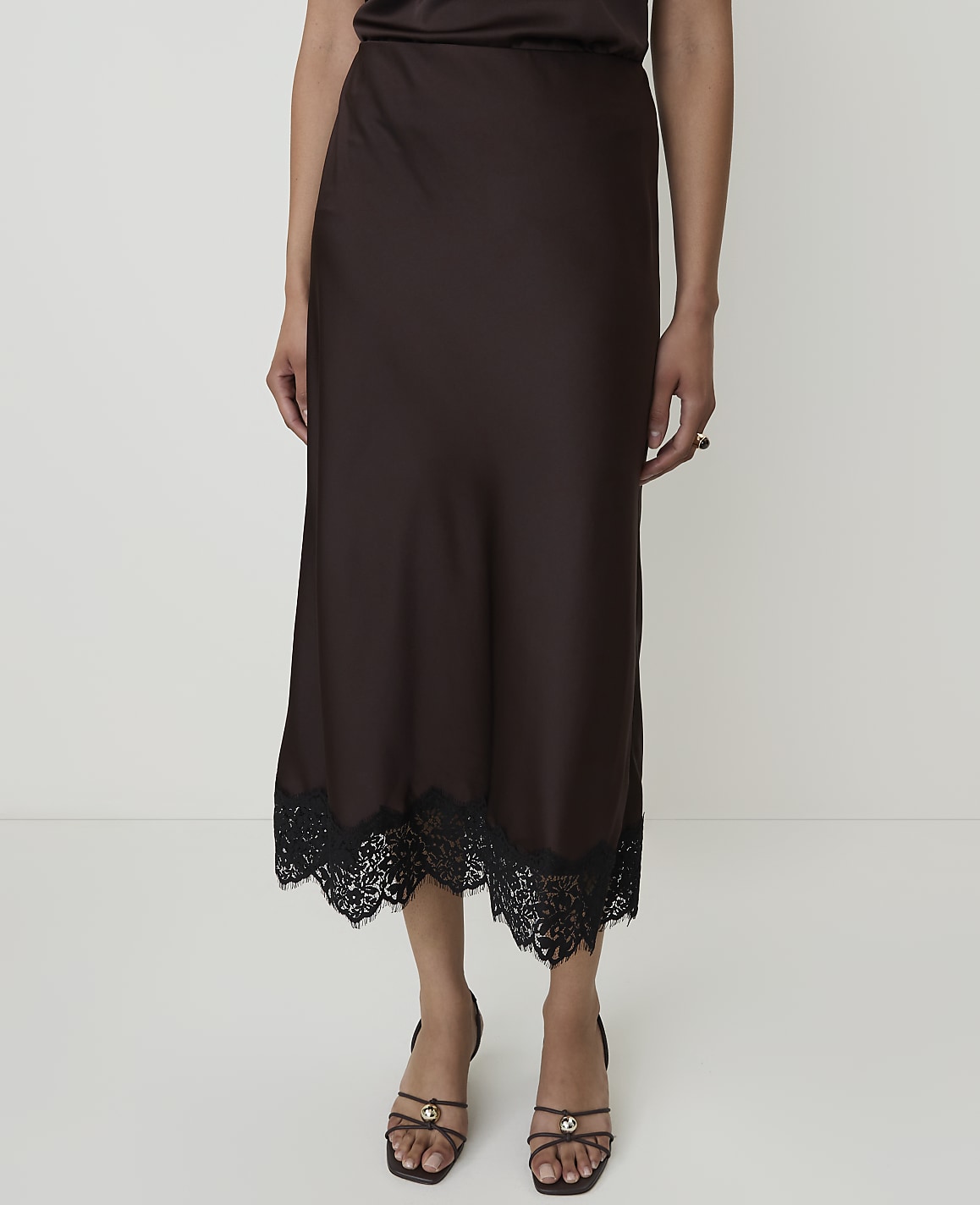 A person stands in front of a plain white background, wearing a brown satin skirt with a black lace trim and strappy sandals.