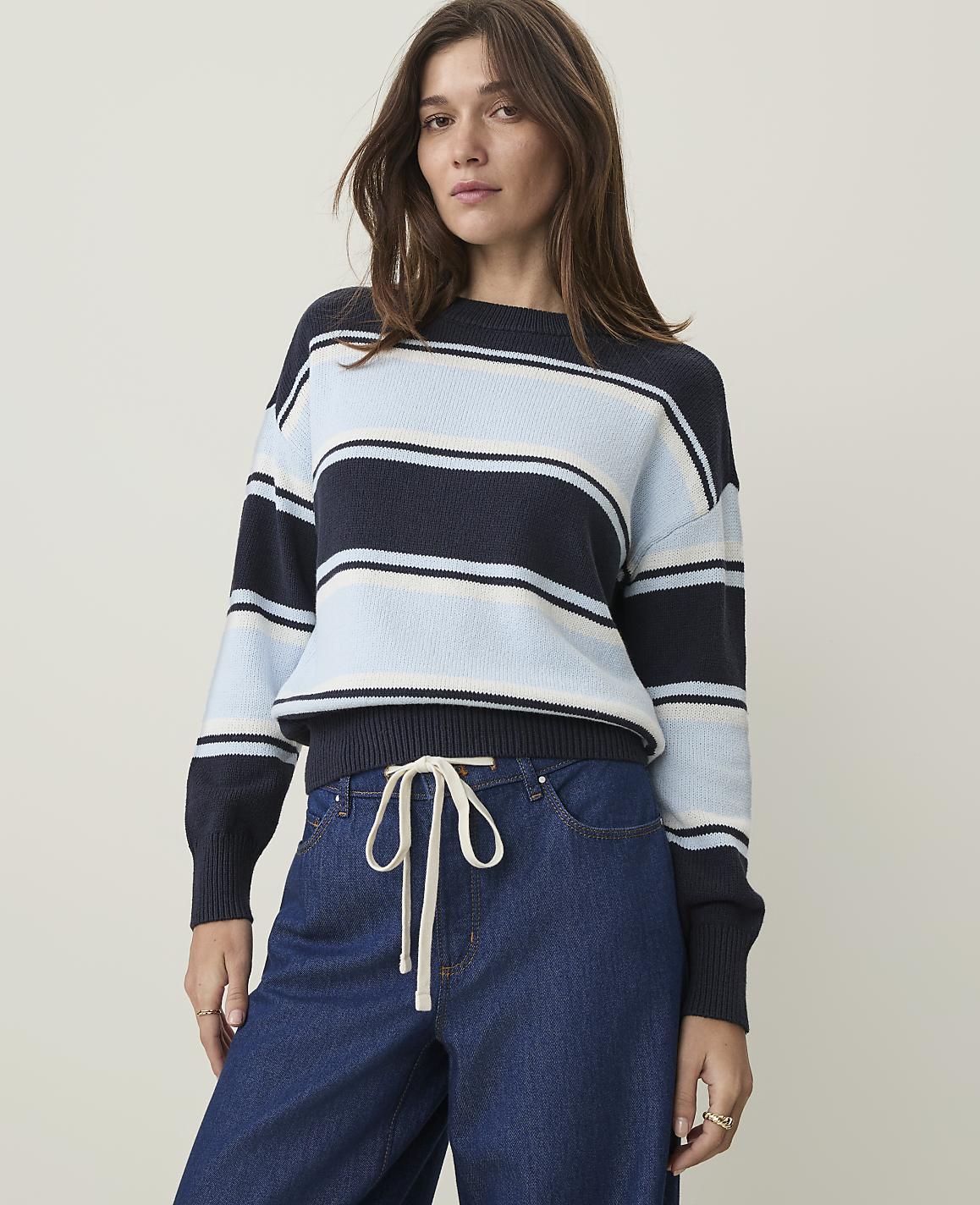 A woman in a striped sweater and jeans stands against a plain, light background.