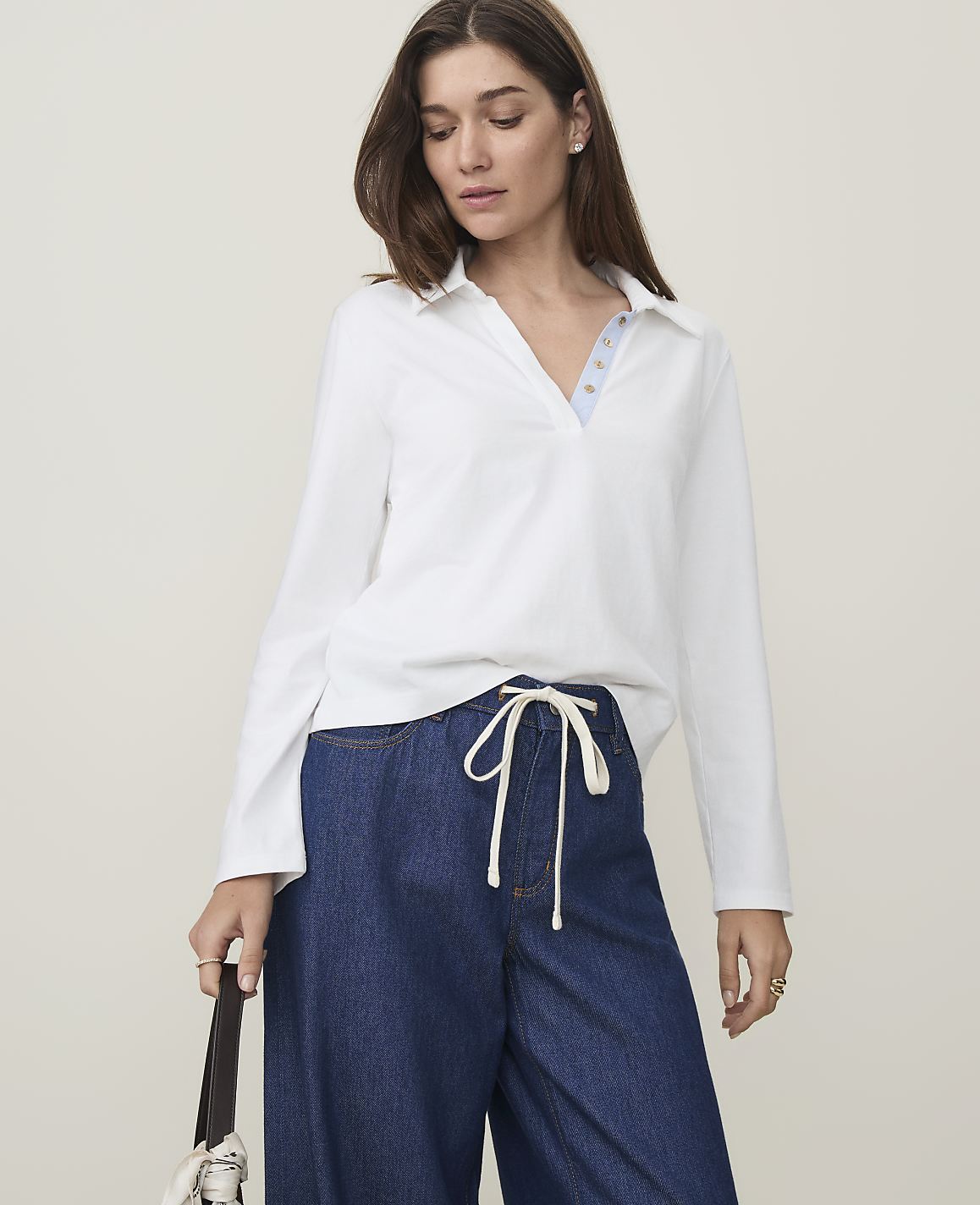 A woman in a white collared shirt and wide-leg jeans stands against a plain, light-colored background.