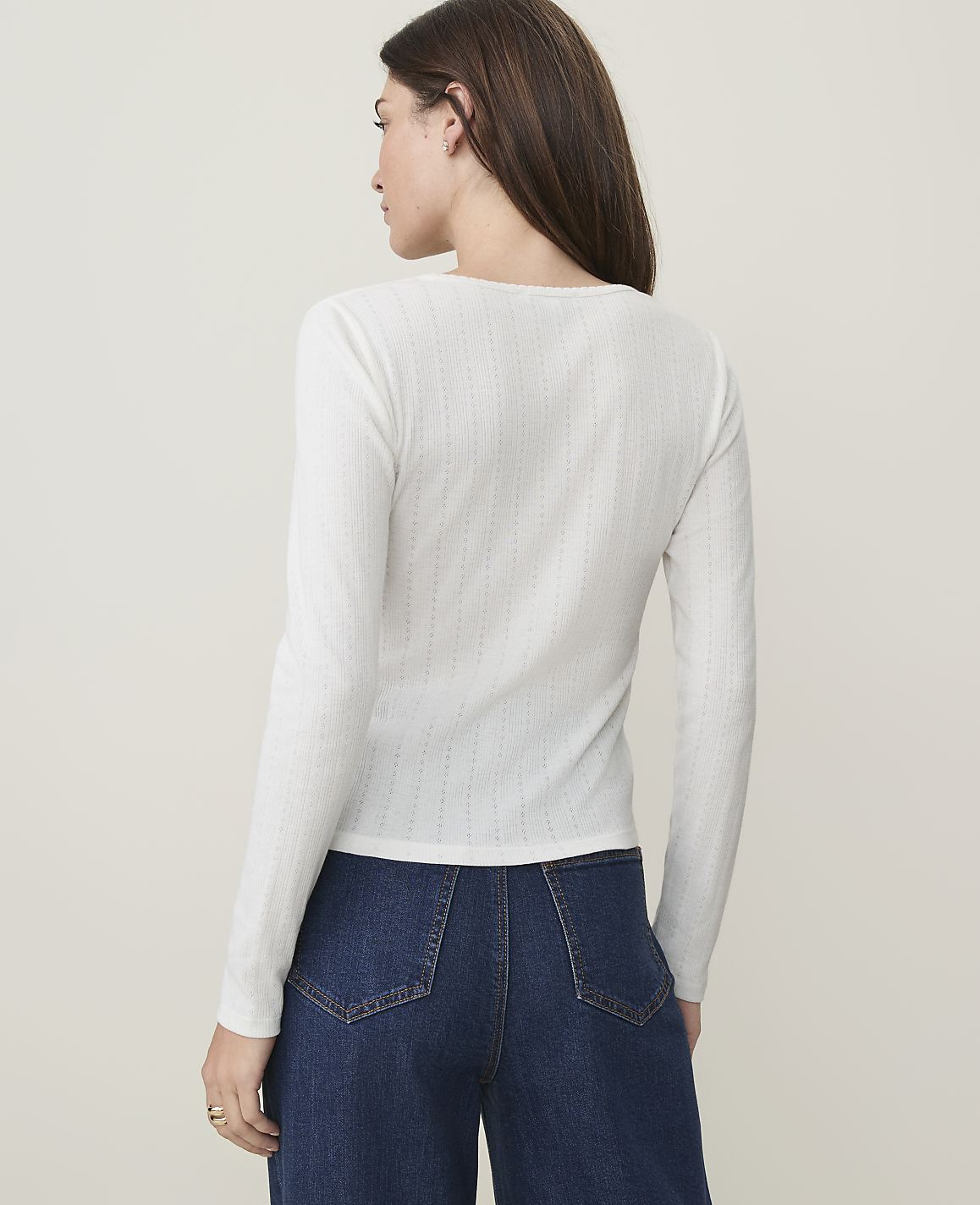 A woman in a white long-sleeved shirt and blue jeans stands against a plain, light-colored background.