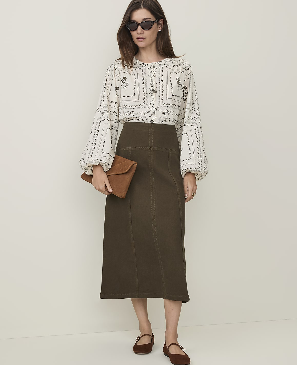 A woman in a patterned blouse and olive green skirt stands against a plain, light-colored background, holding a brown clutch.