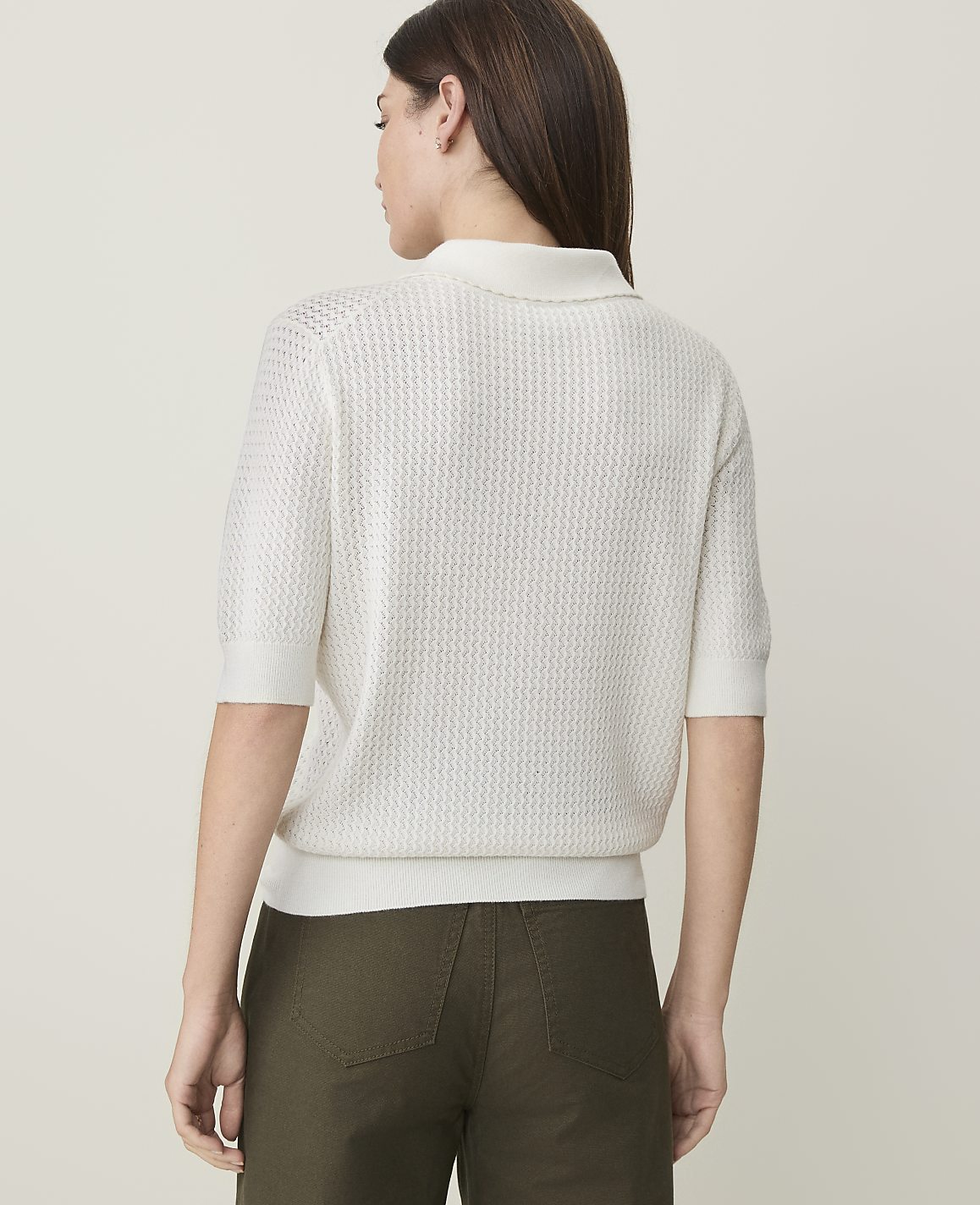 A woman in a white knit polo shirt and olive green pants stands against a plain white background.