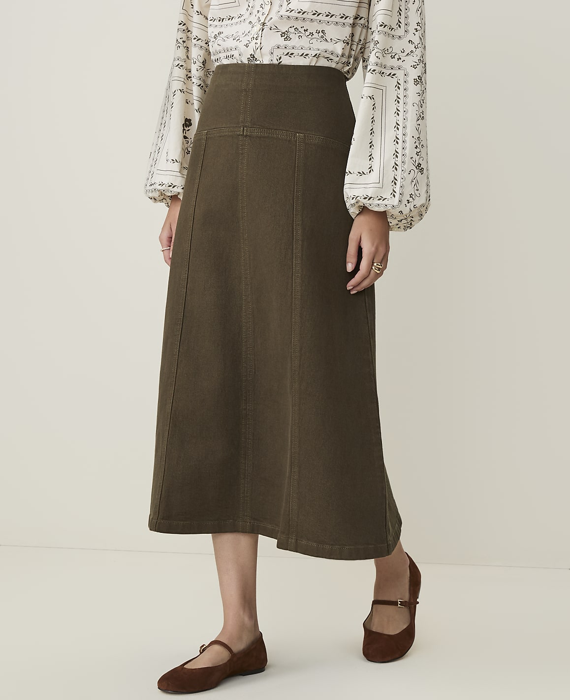 A woman in a patterned blouse and a long olive-green skirt stands against a plain, light-colored background.